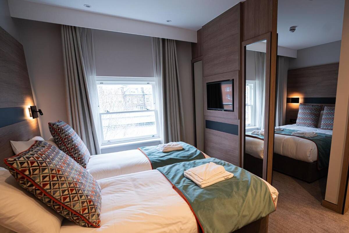 A hotel room with two beds, a large window, a mirror, and a television.