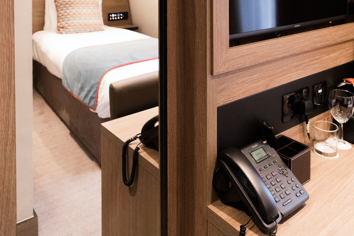 A hotel room with a bed, a mounted TV, a telephone, and a glass of wine on the nightstand.