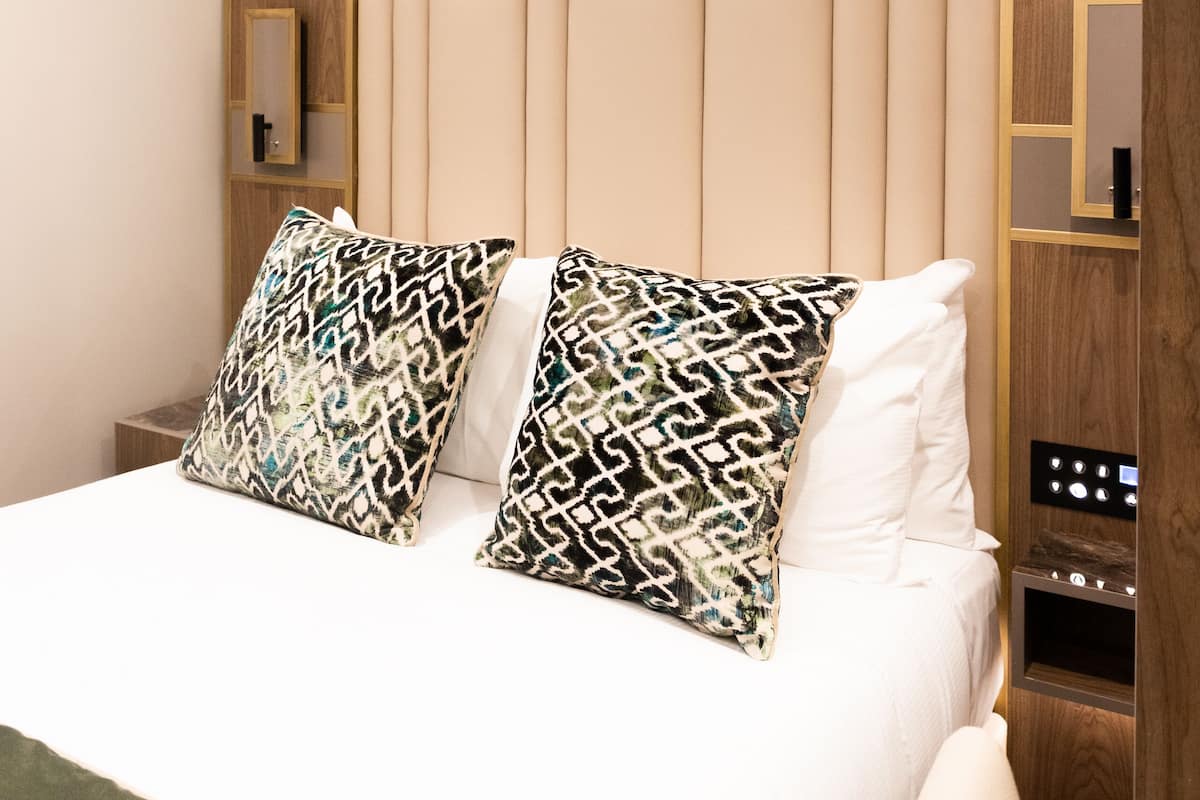 A modern hotel room with a wooden headboard, two patterned pillows, a white bedspread, and a bedside table with a control panel.