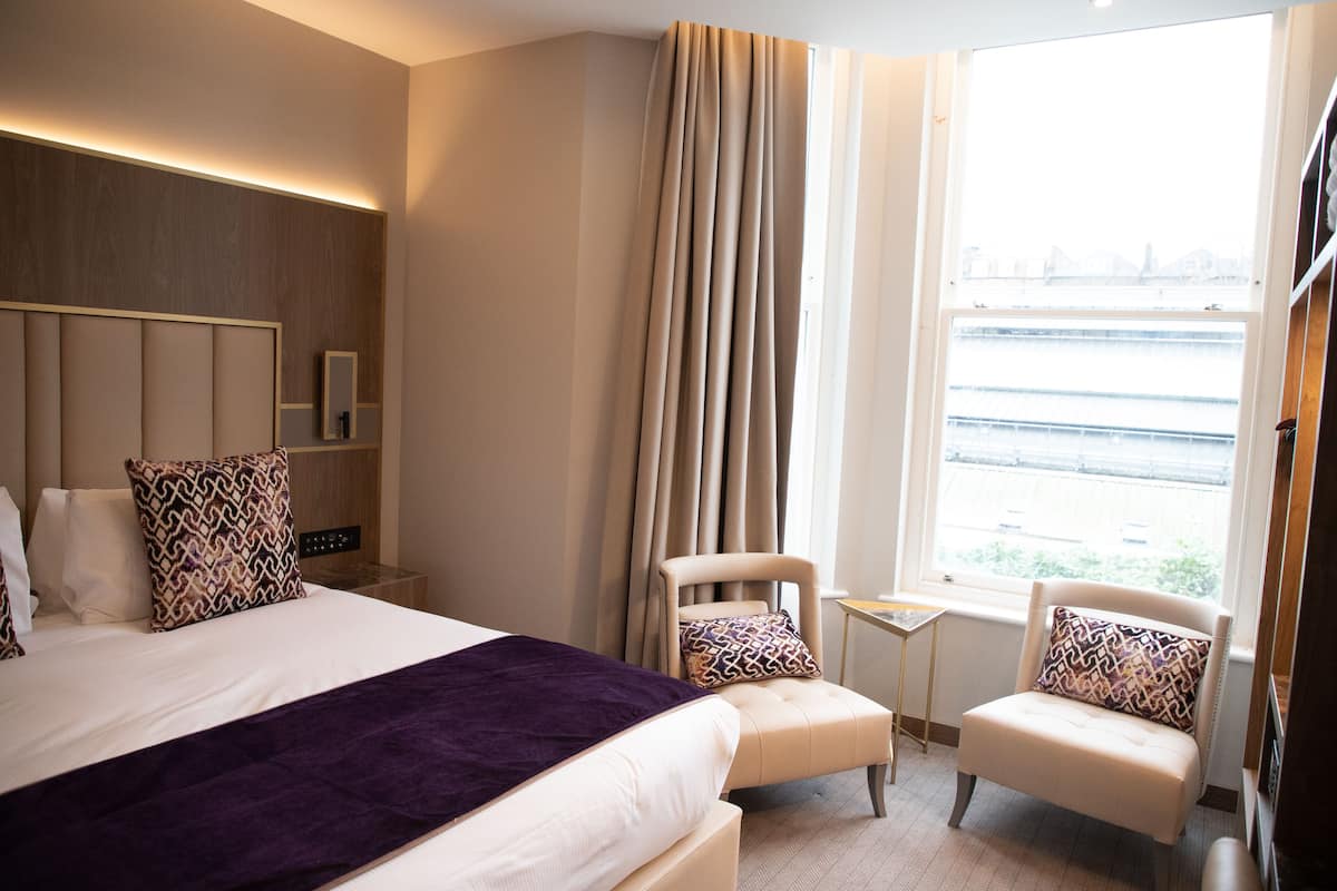 A hotel room with a large bed, two armchairs, a small table, a mirror, and a window with a view of a bridge.