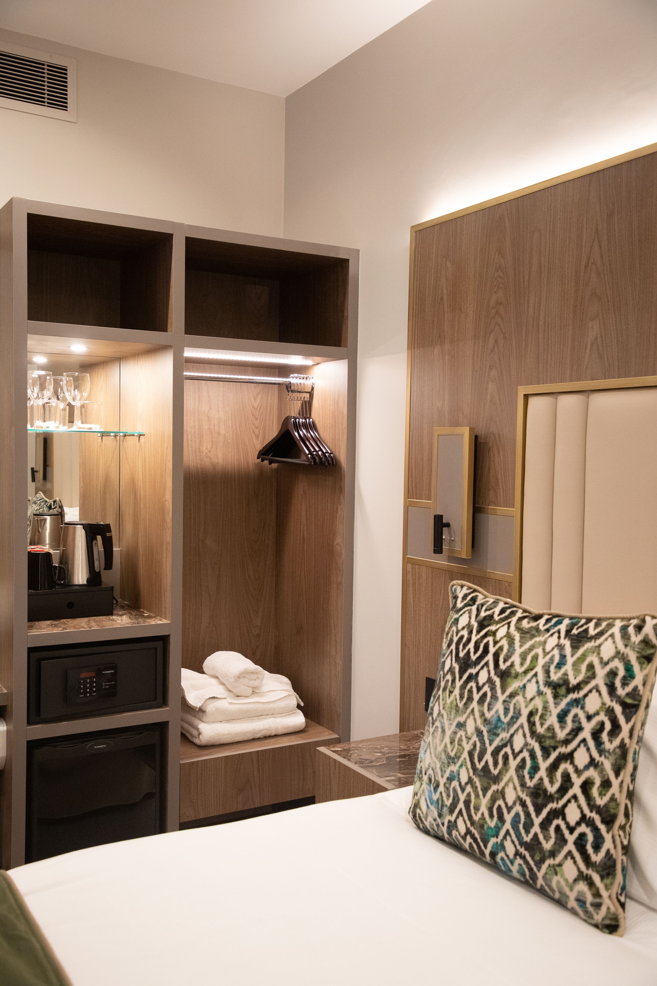 A hotel room with a bed, a wardrobe with built-in shelves, a mirror, and a bedside table with a lamp.