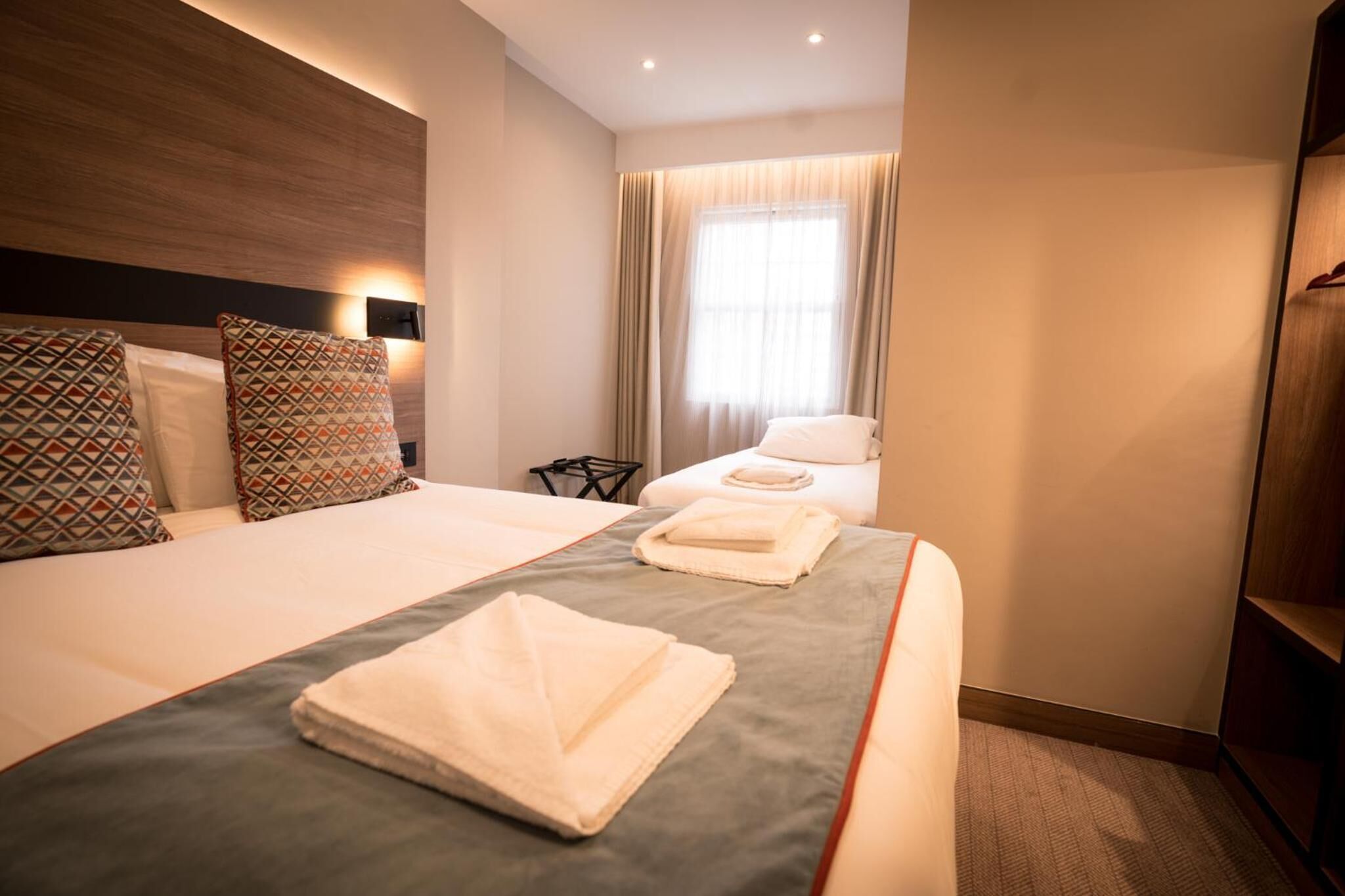 A hotel room with two beds, a wooden headboard, a bedside table, a lamp, and a window with curtains.