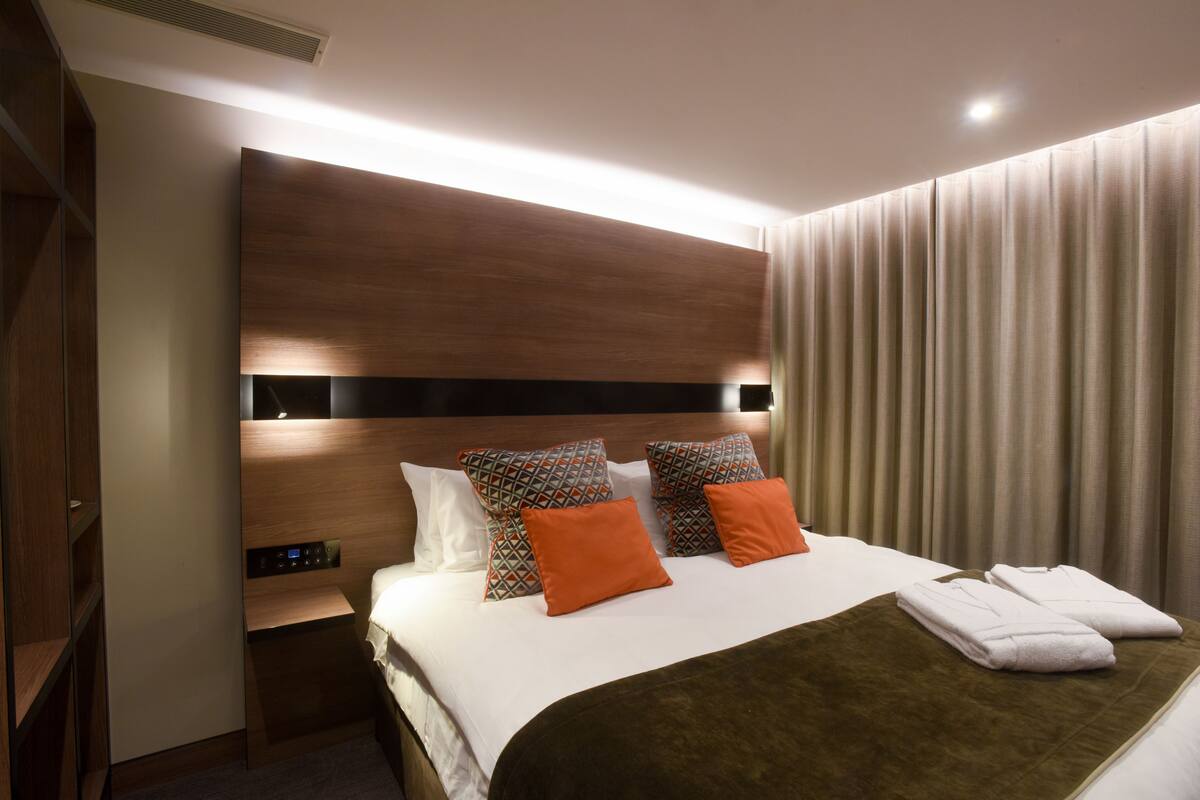 A modern hotel room with a large bed, a wooden headboard, and a control panel on the side.