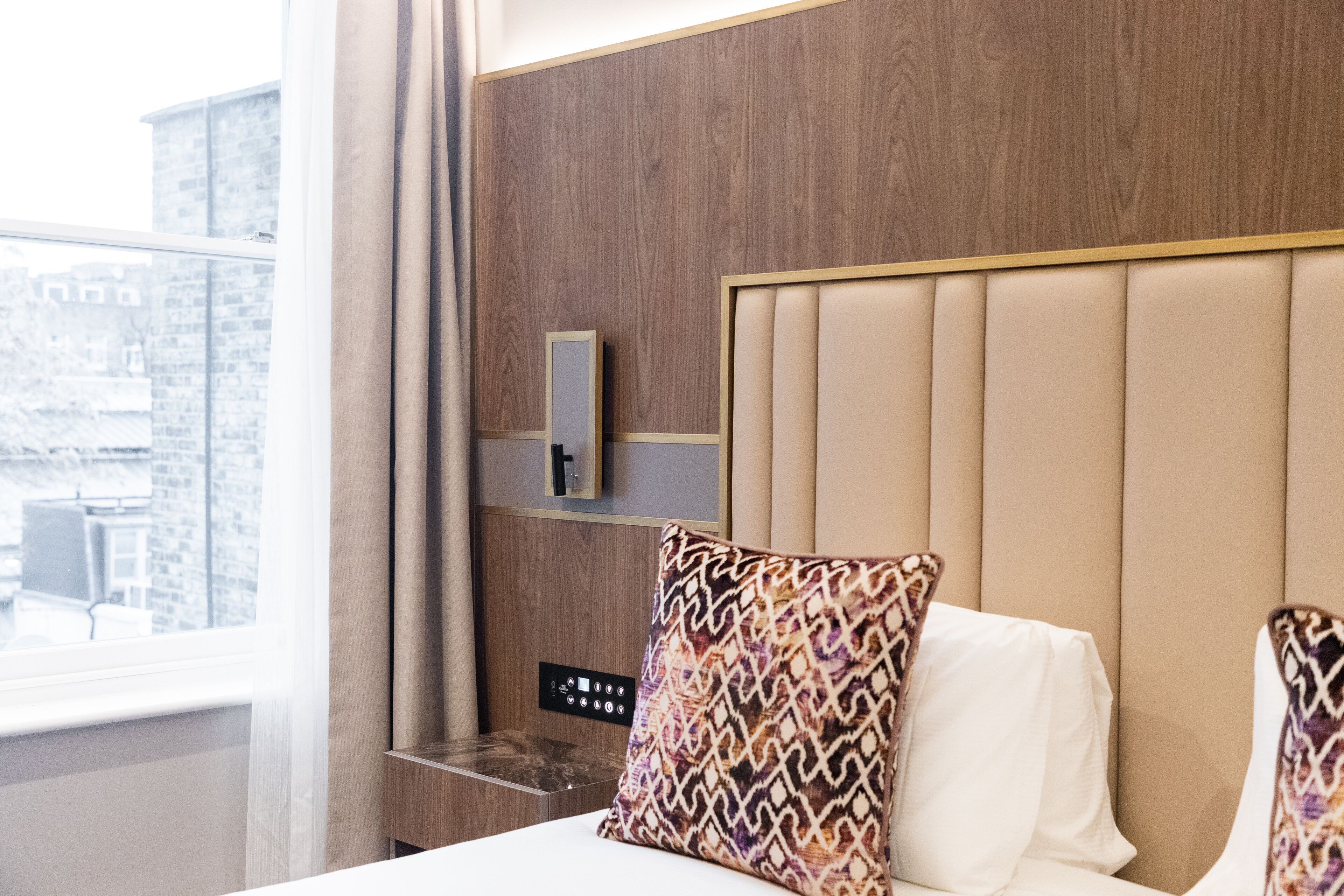 A hotel room with a wooden headboard, a bedside table, and a window with curtains.