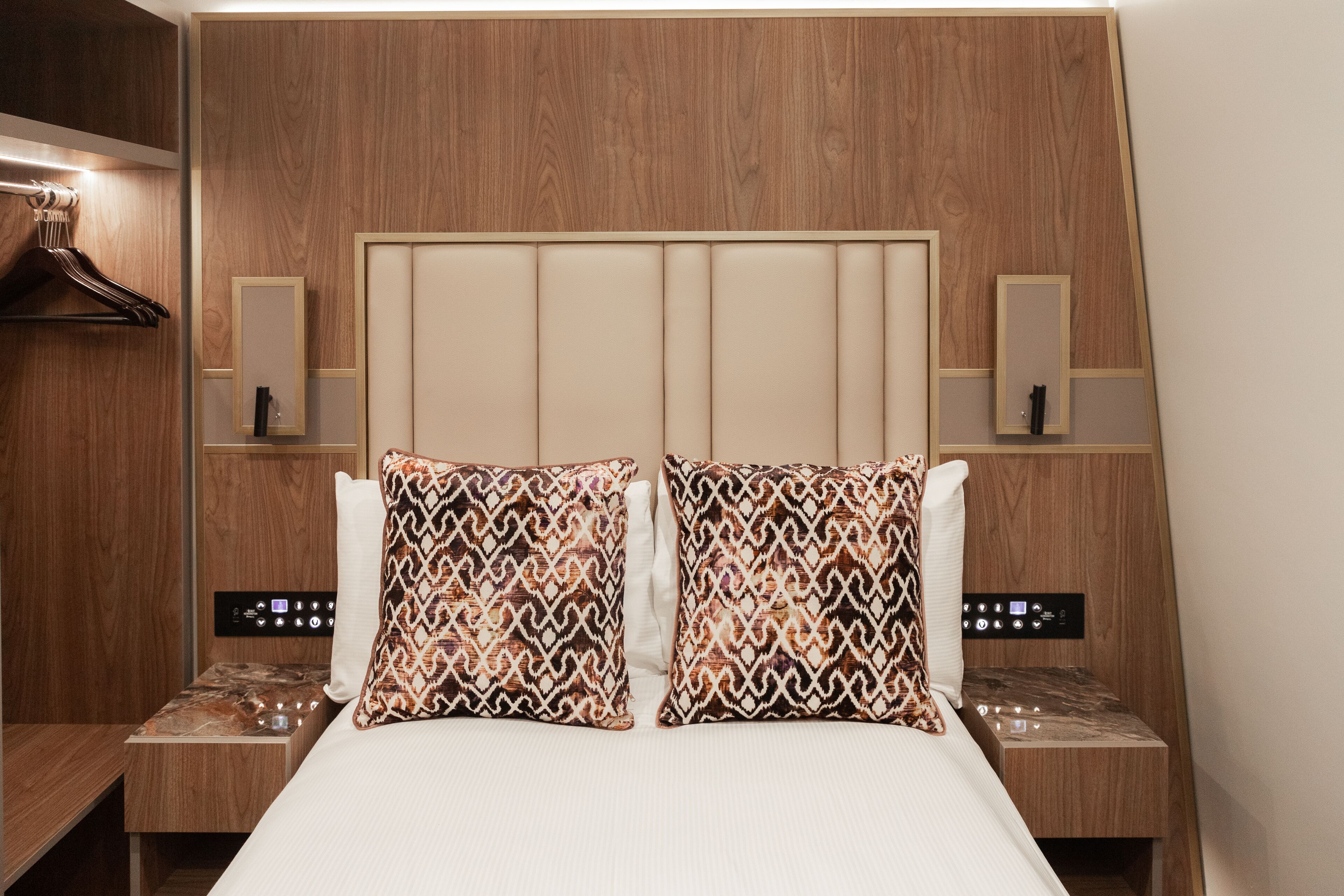 Habitación de hotel con cabecera de madera, dos almohadas estampadas, una mesita de noche con panel de control y un perchero.