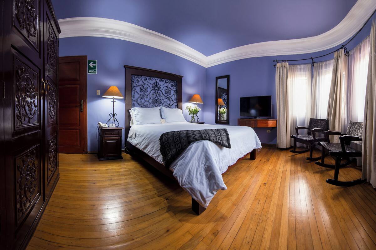 A bedroom with a large bed, wooden floor, a TV, and a desk with chairs.