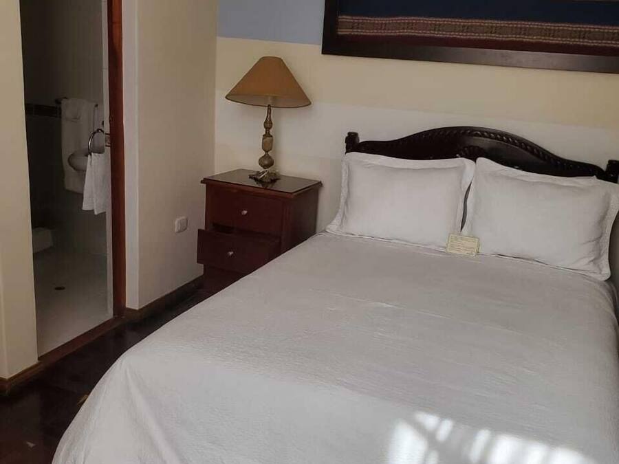 Una cama bien hecha con sábanas blancas, una mesita de noche de madera con una lámpara, y un cuadro en la pared.
