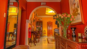 A hallway with a red wall, a large mirror, a bench, a chandelier, and a vase with flowers. - Casa Arequipa (Arequipa)