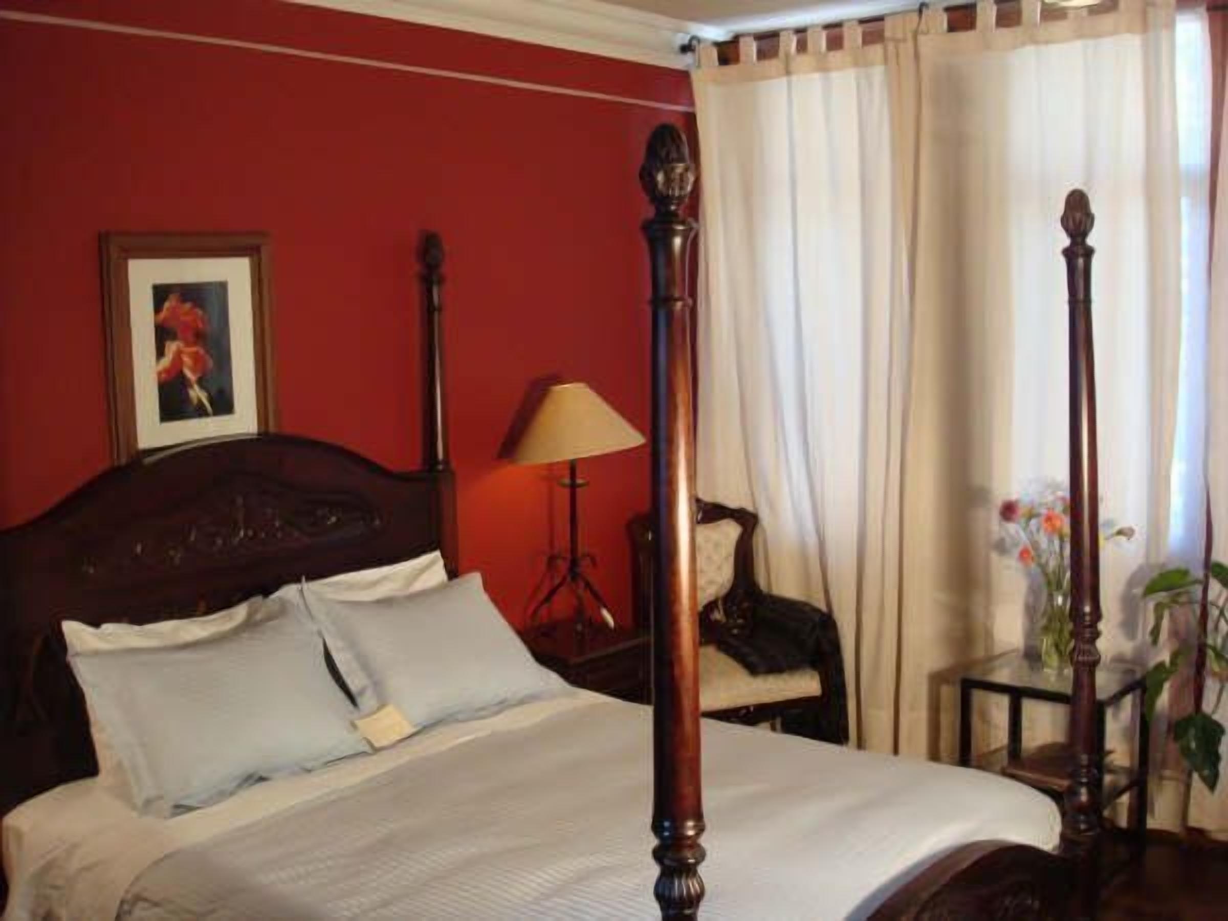 A four-poster bed with white linens, a red wall, a framed picture, a lamp, and a chair.