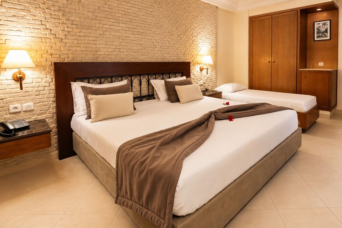 Una camera d'albergo con un letto grande, un letto più piccolo, un armadio in legno, un comodino con un telefono e una parete in mattoni.