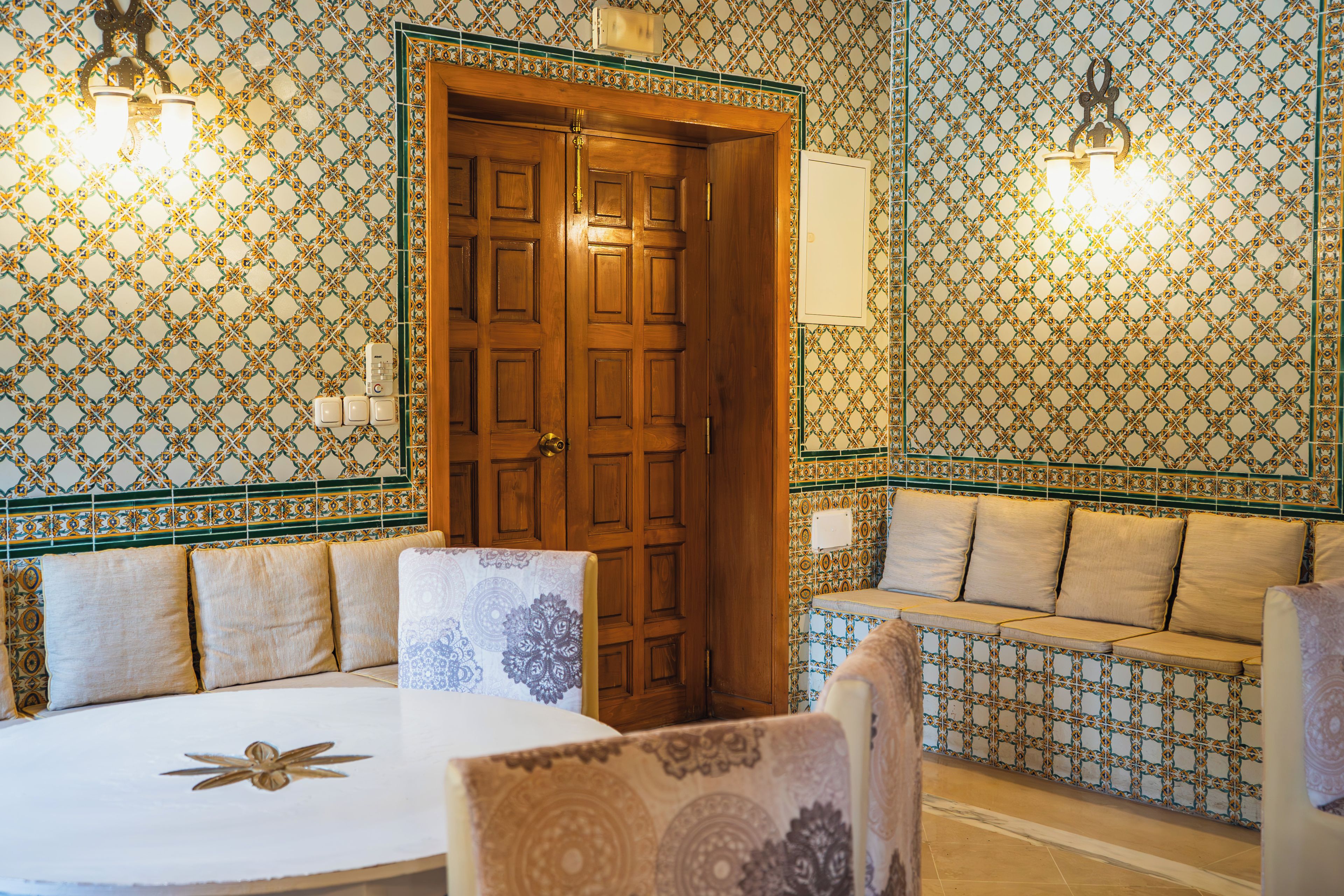 A room with a wooden door, patterned walls, a round table, and a sofa with cushions.