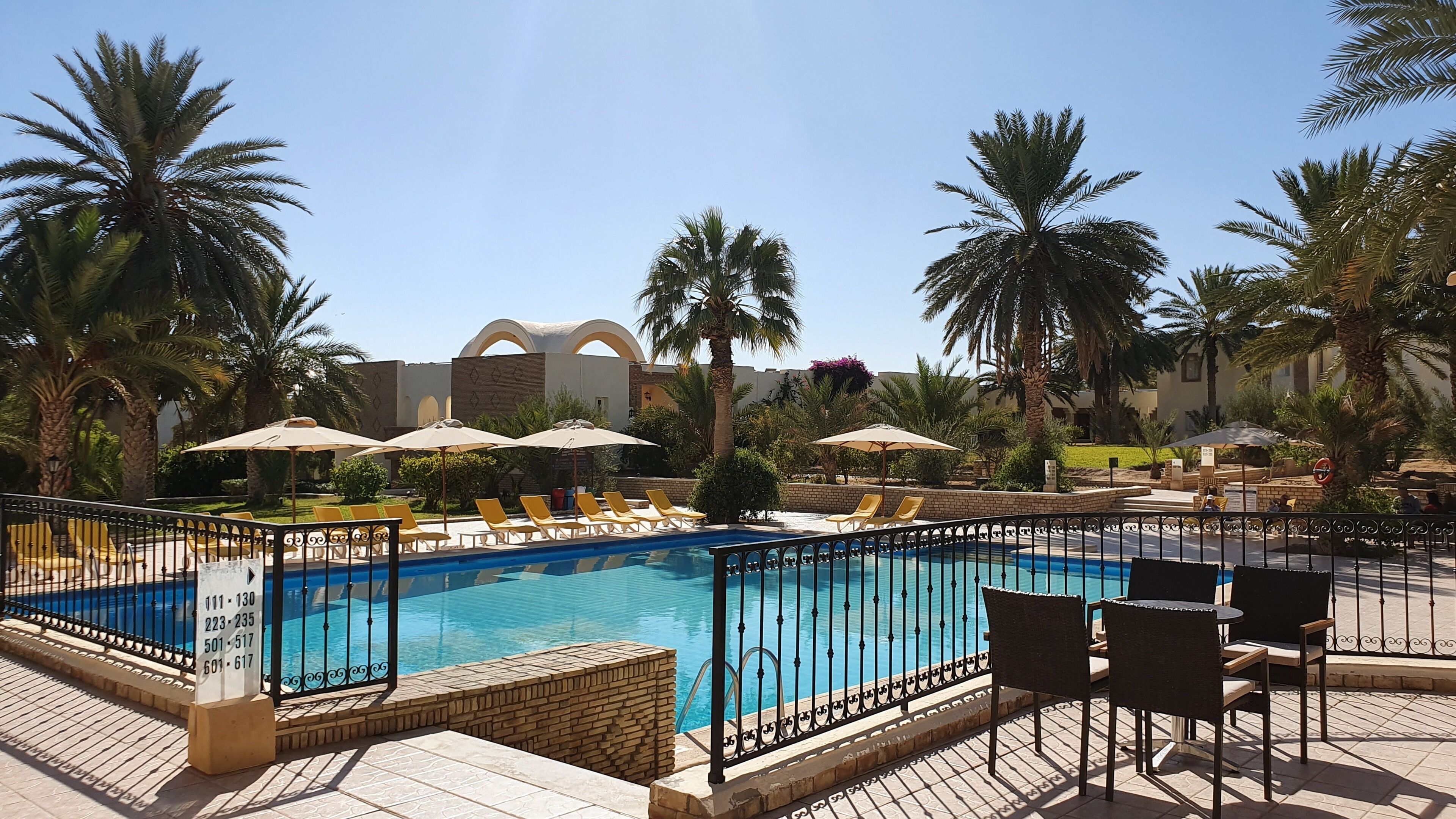 A pool area with lounge chairs and umbrellas, surrounded by palm trees and a building with arches in the background.