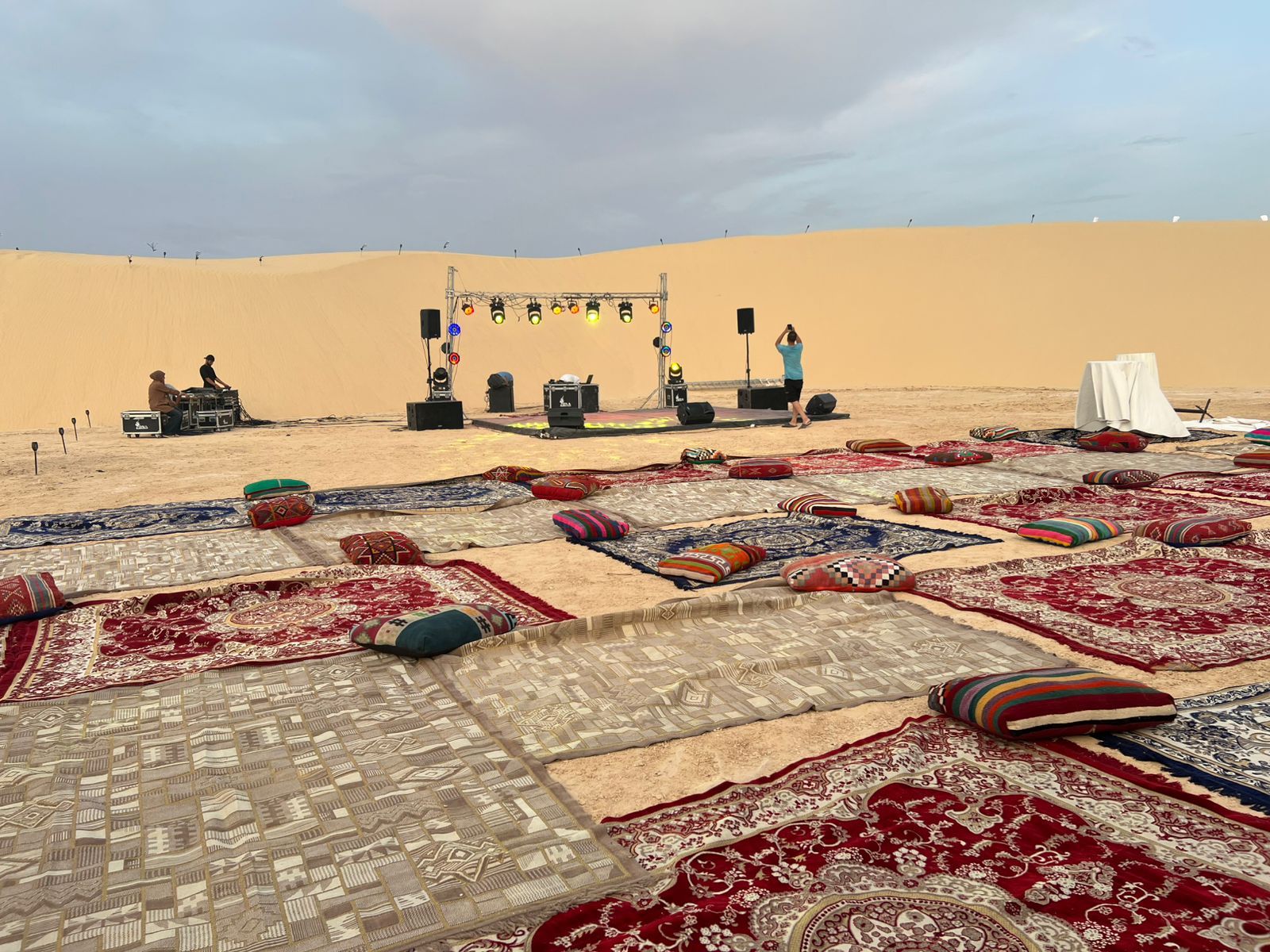 Un paisaje desértico con un escenario y equipo musical, rodeado de alfombras y cojines variados.