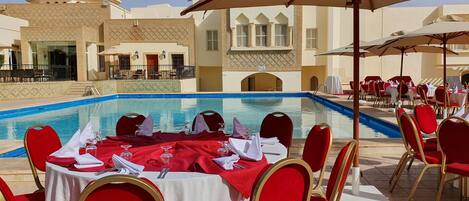 A poolside dining area with tables set for service, red chairs, and umbrellas.