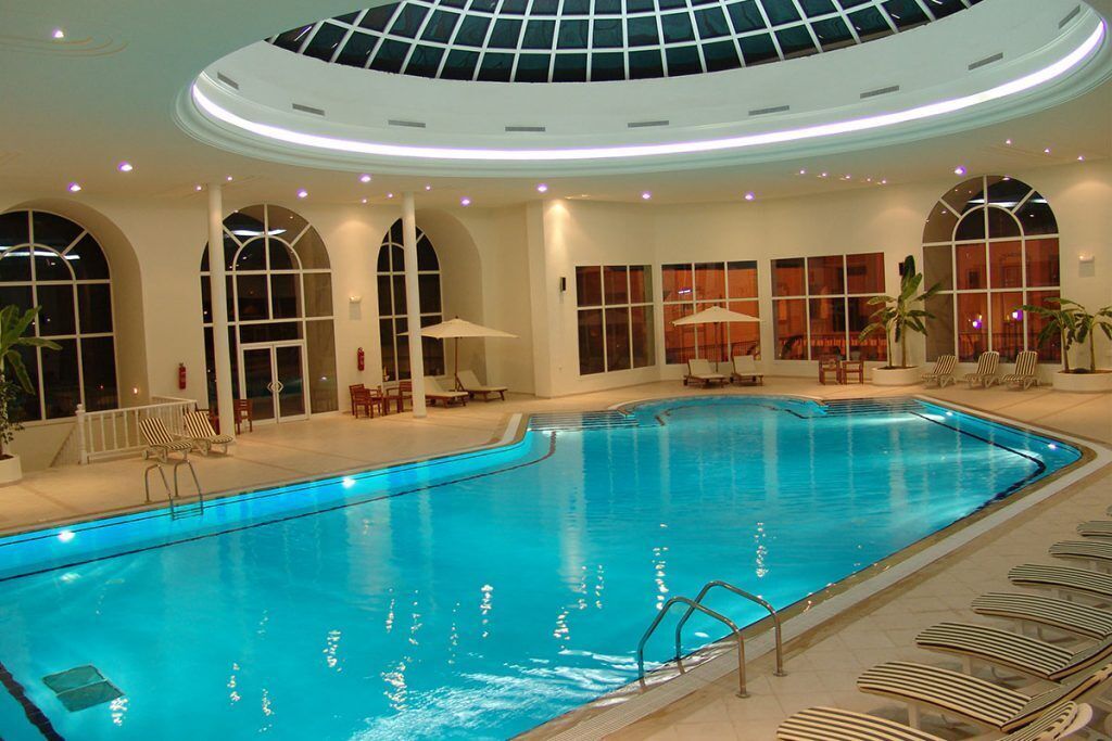 An indoor swimming pool with a glass ceiling, lounge chairs, and sunbeds around the pool area.