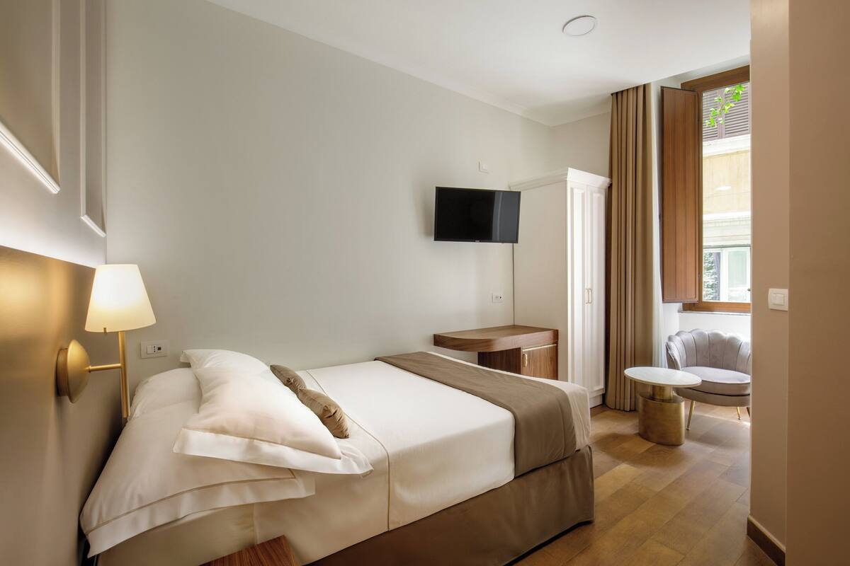 Una camera d'albergo con un letto, una lampada da comodino, una TV, un tavolino e un piccolo tavolo rotondo con una sedia.
