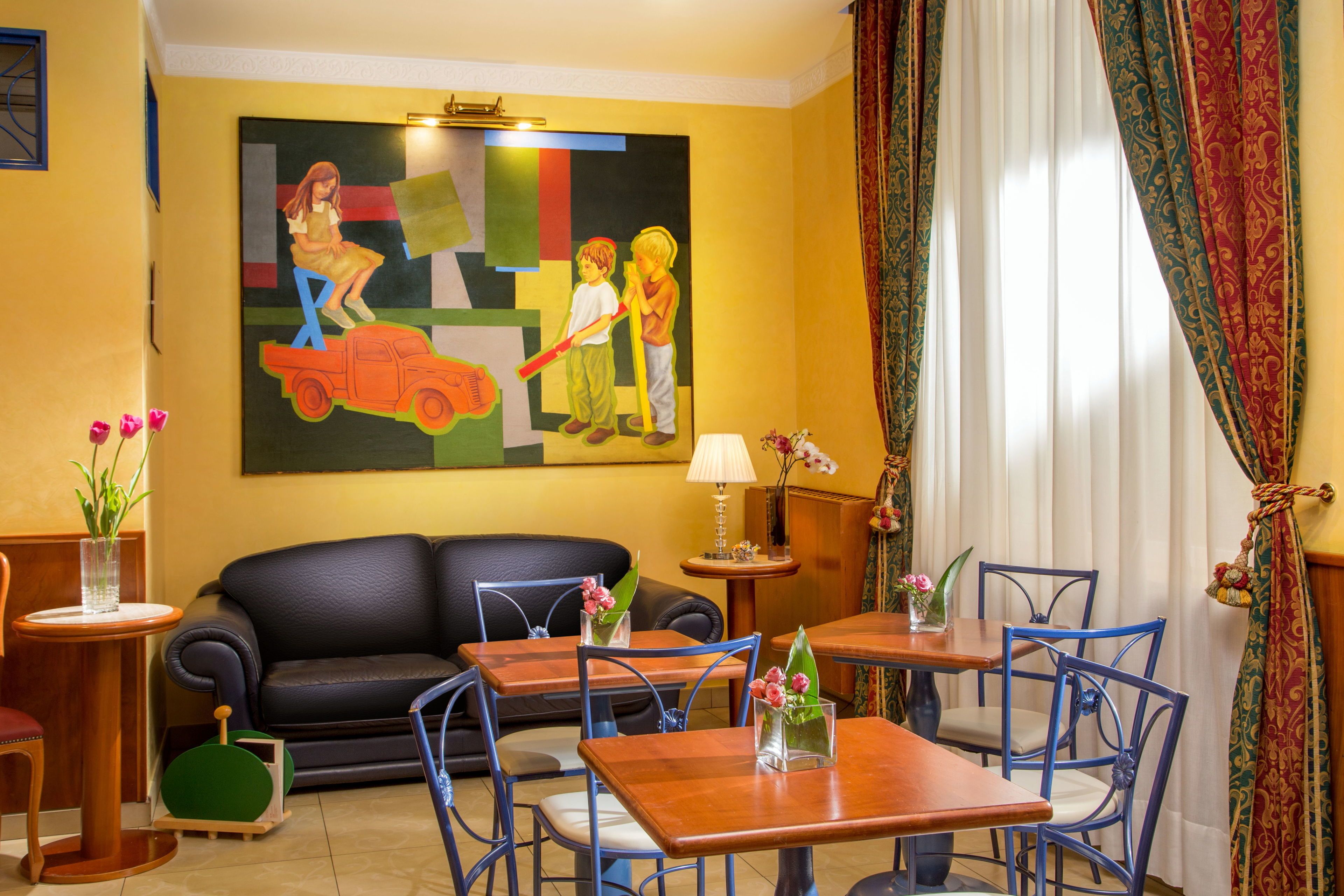 Quarto de hotel com sofá de couro preto, mesas de madeira, cadeiras azuis, um quadro na parede e uma janela com cortinas.
