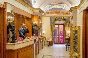 Una zona de recepción de hotel con mostrador de mármol, un espejo grande, una estatua y un techo decorativo.