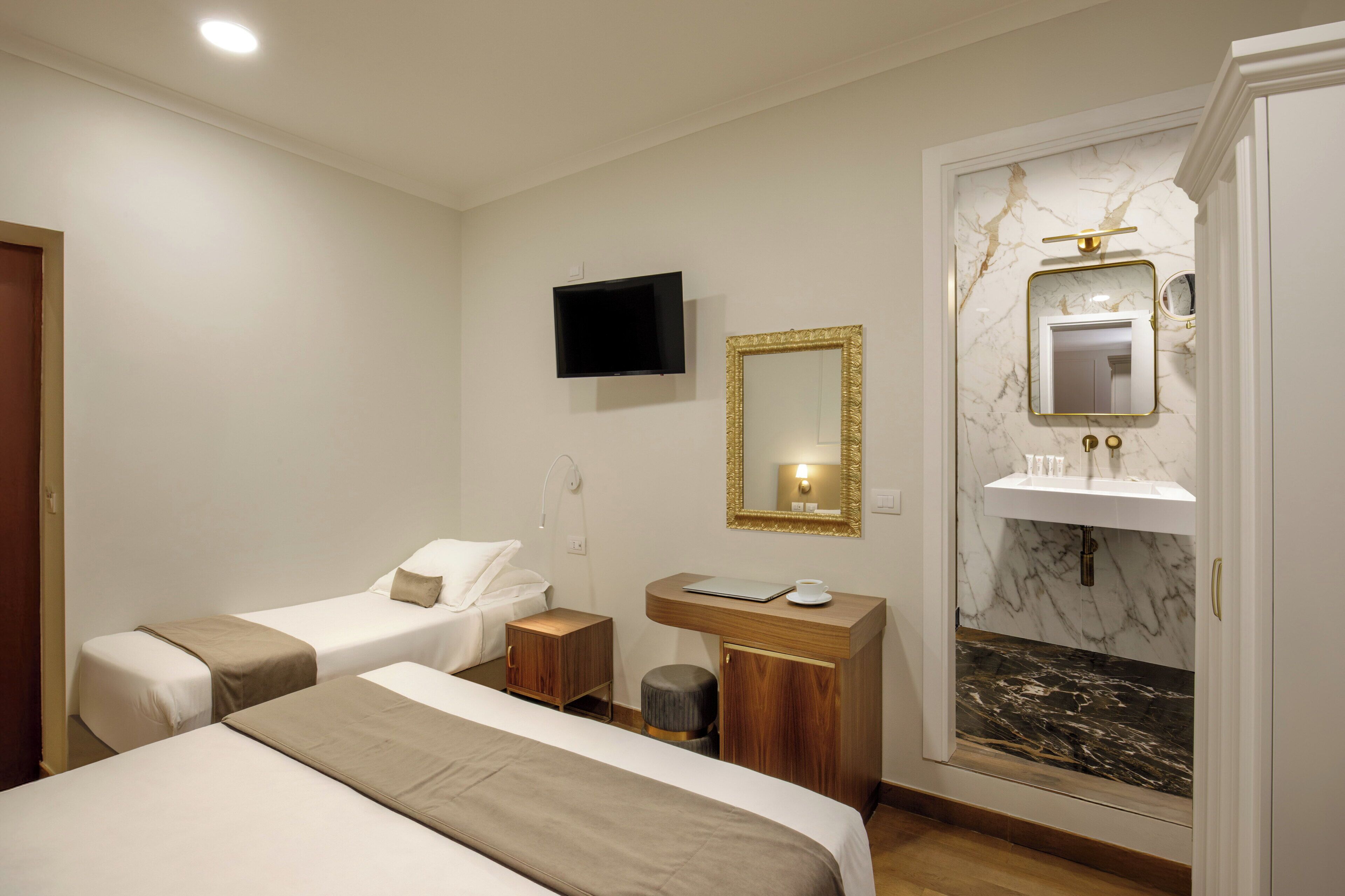 A hotel room with two beds, a TV, a vanity with a mirror, and a marble sink.