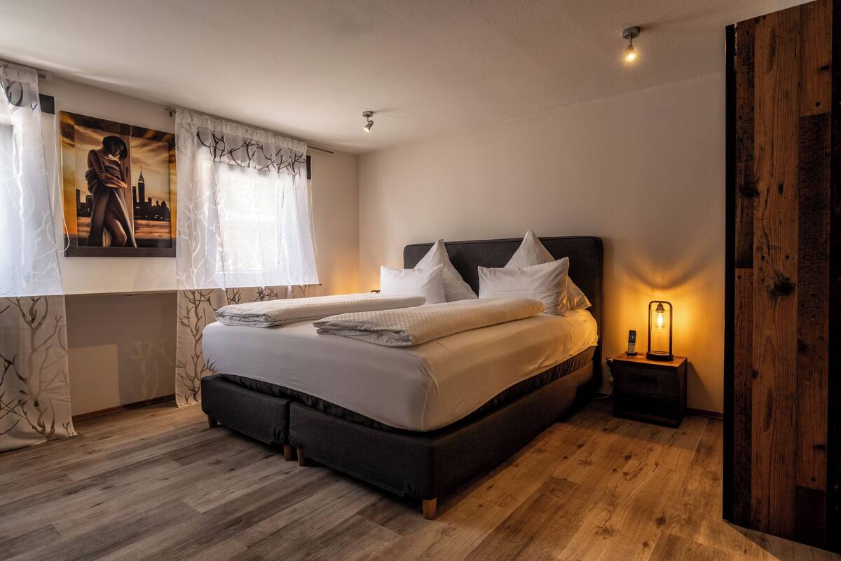 A bedroom with a large bed, a bedside table with a lamp, and a cityscape painting on the wall.