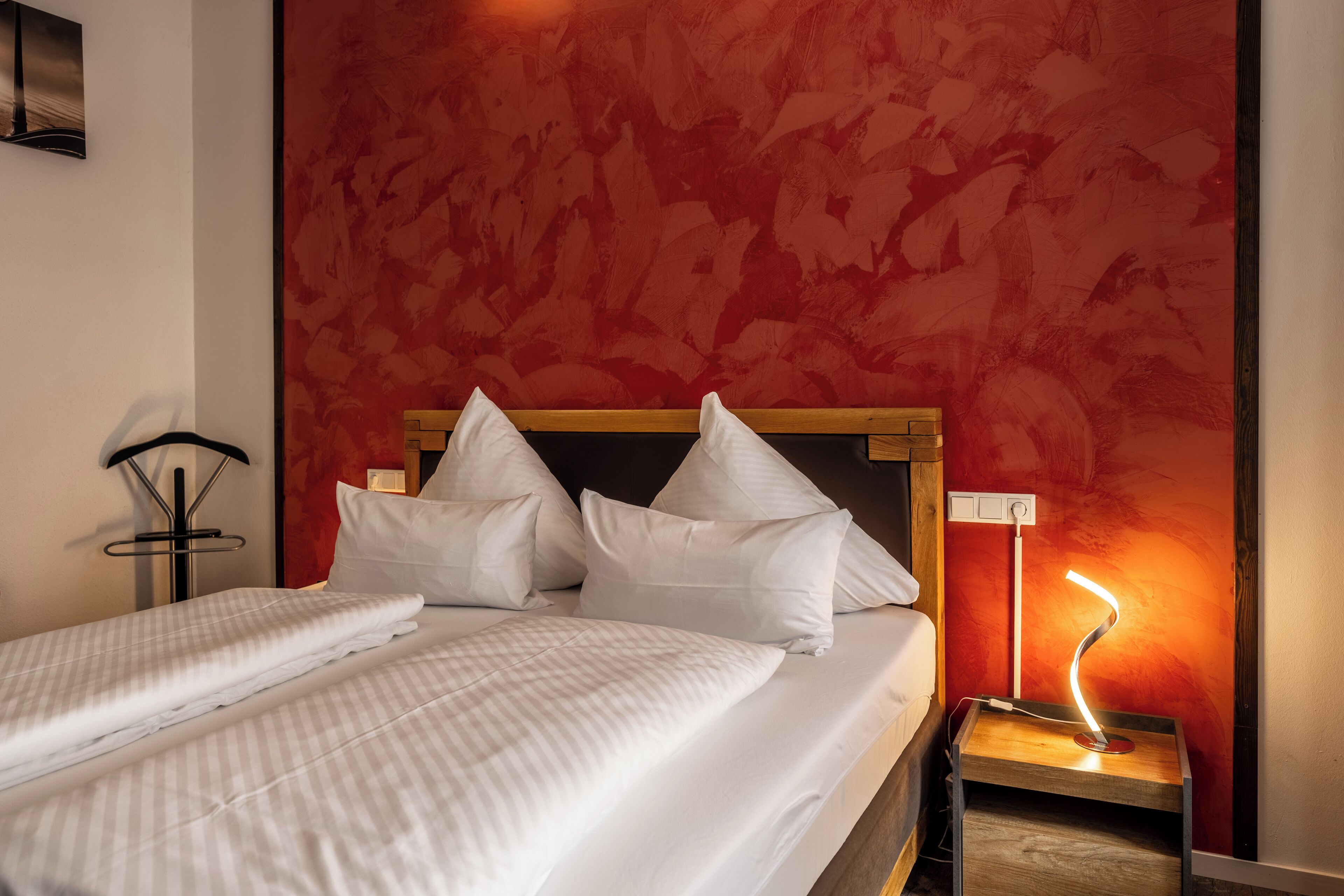A neatly made bed with white linens and pillows, a bedside lamp, and a red textured wall.