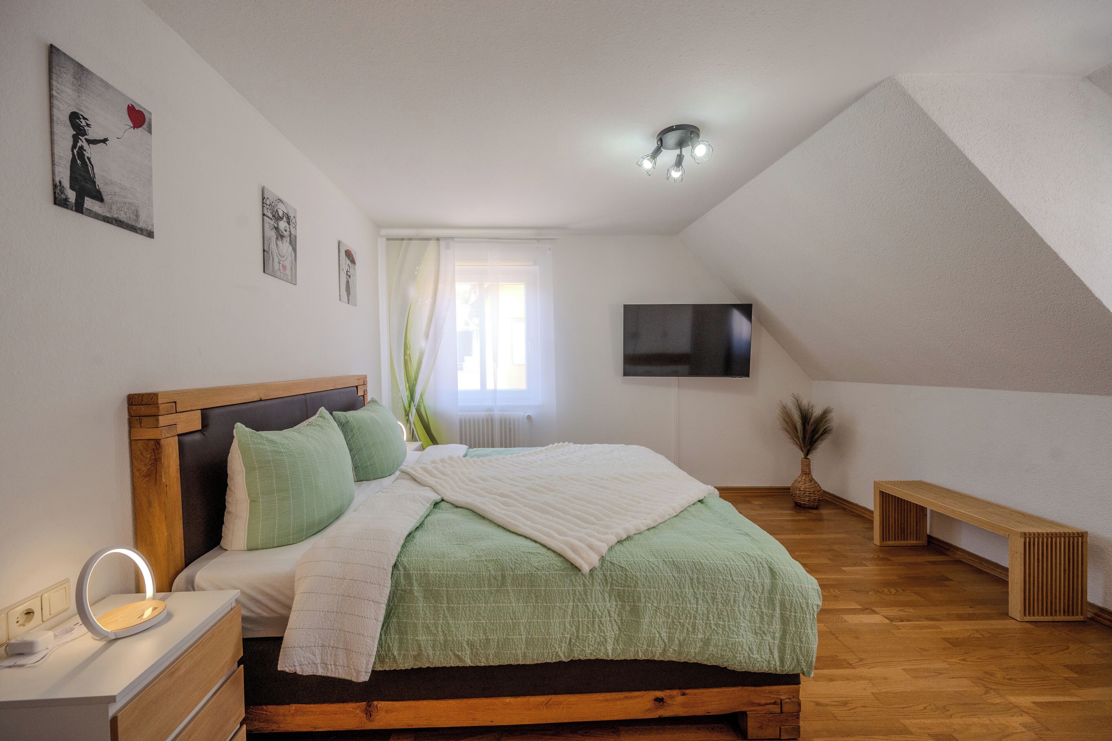 A bedroom with a bed, bedside table, lamp, wall-mounted TV, and a window with curtains.
