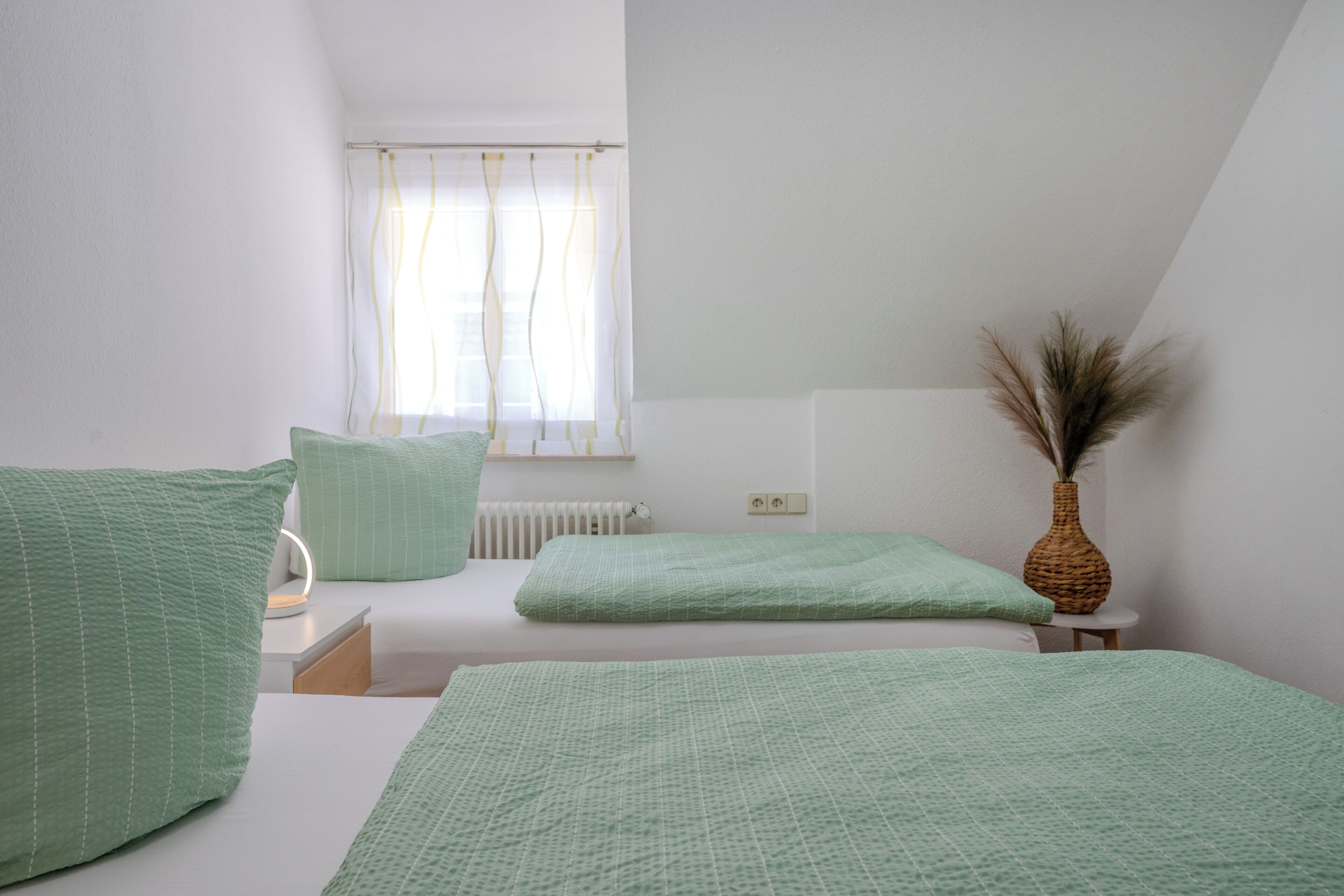 A bedroom with two beds, a radiator, a window with curtains, and a vase with dried plants.