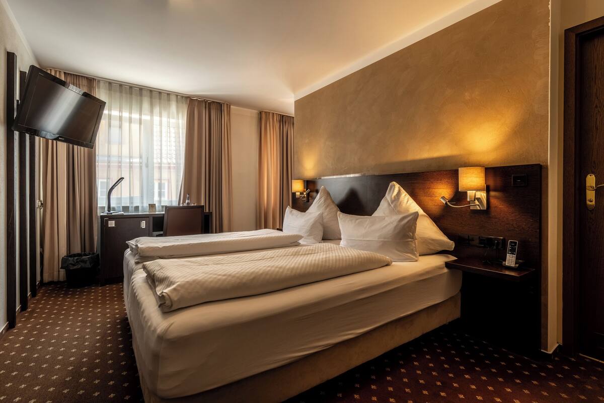 A hotel room with two beds, a TV mounted on the wall, a desk, and a nightstand with a lamp.