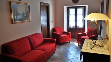 Un salon avec un canapé rouge, deux fauteuils rouges, une table avec une lampe, un tableau au mur et une porte avec une fenêtre.