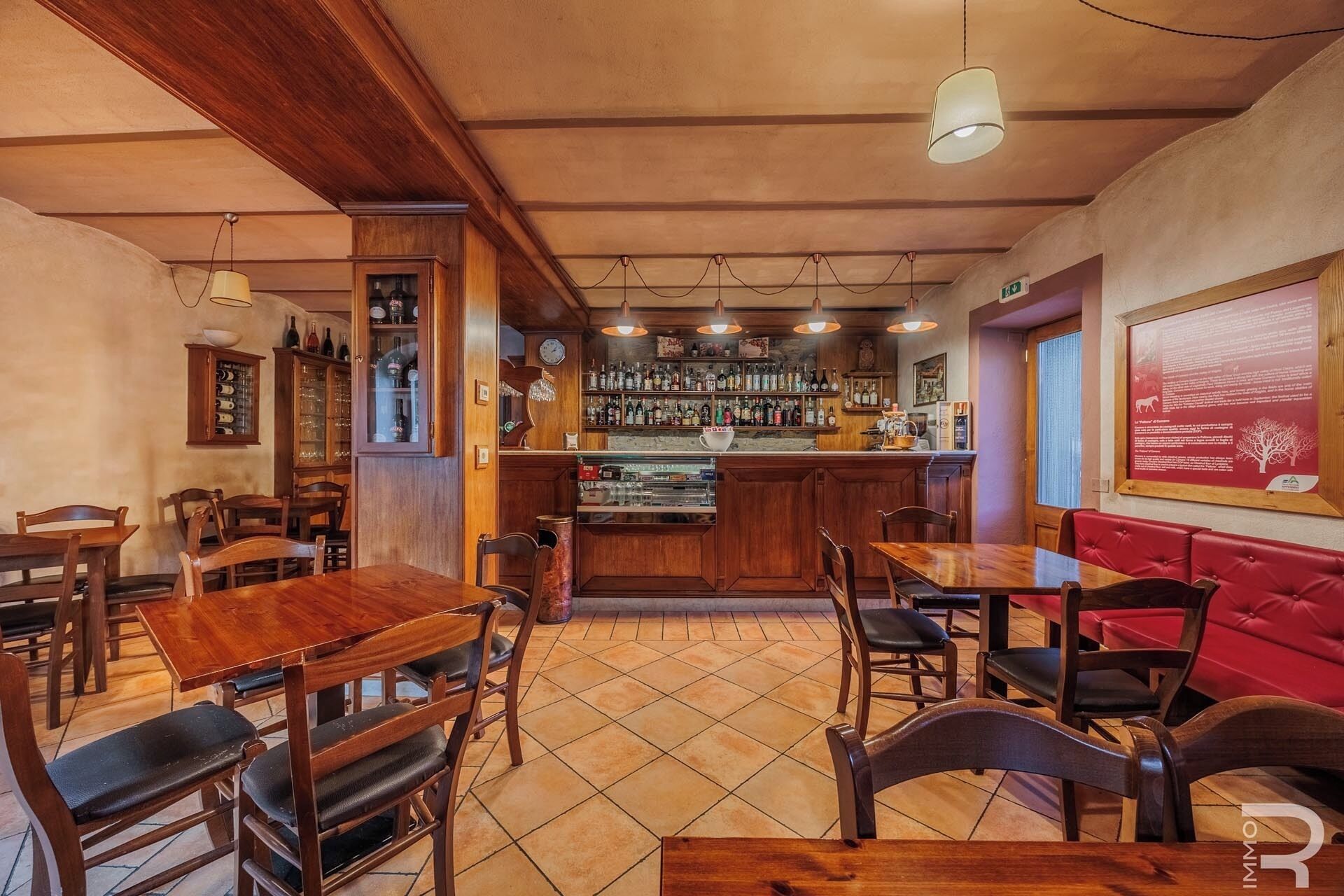Un restaurant avec des tables et des chaises en bois, un bar avec des étagères remplies de bouteilles et un coin salon avec des banquettes rouges.