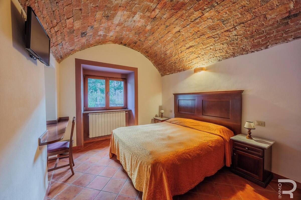Ein Schlafzimmer mit Ziegeldecke, einem hölzernen Bett, einem Nachttisch, einem Sessel und einem Fenster mit Blick auf Bäume.