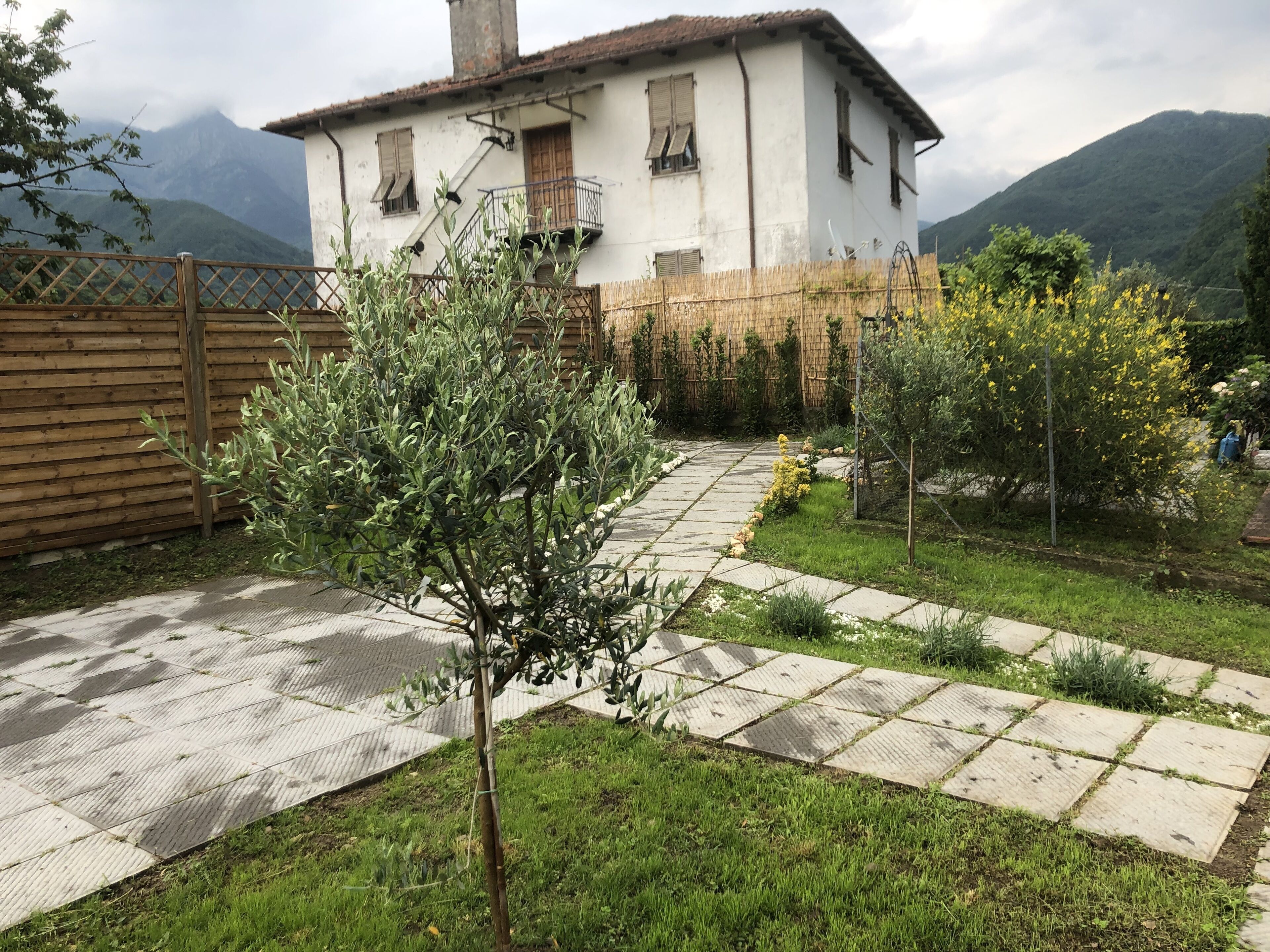 Una casa blanca con balcón, rodeada de un jardín con un sendero empedrado, cerca de una valla de madera y con una montaña al fondo.