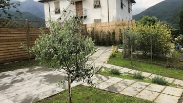 Una casa blanca con balcón, rodeada de un jardín con un sendero empedrado, cerca de una valla de madera y con una montaña al fondo.