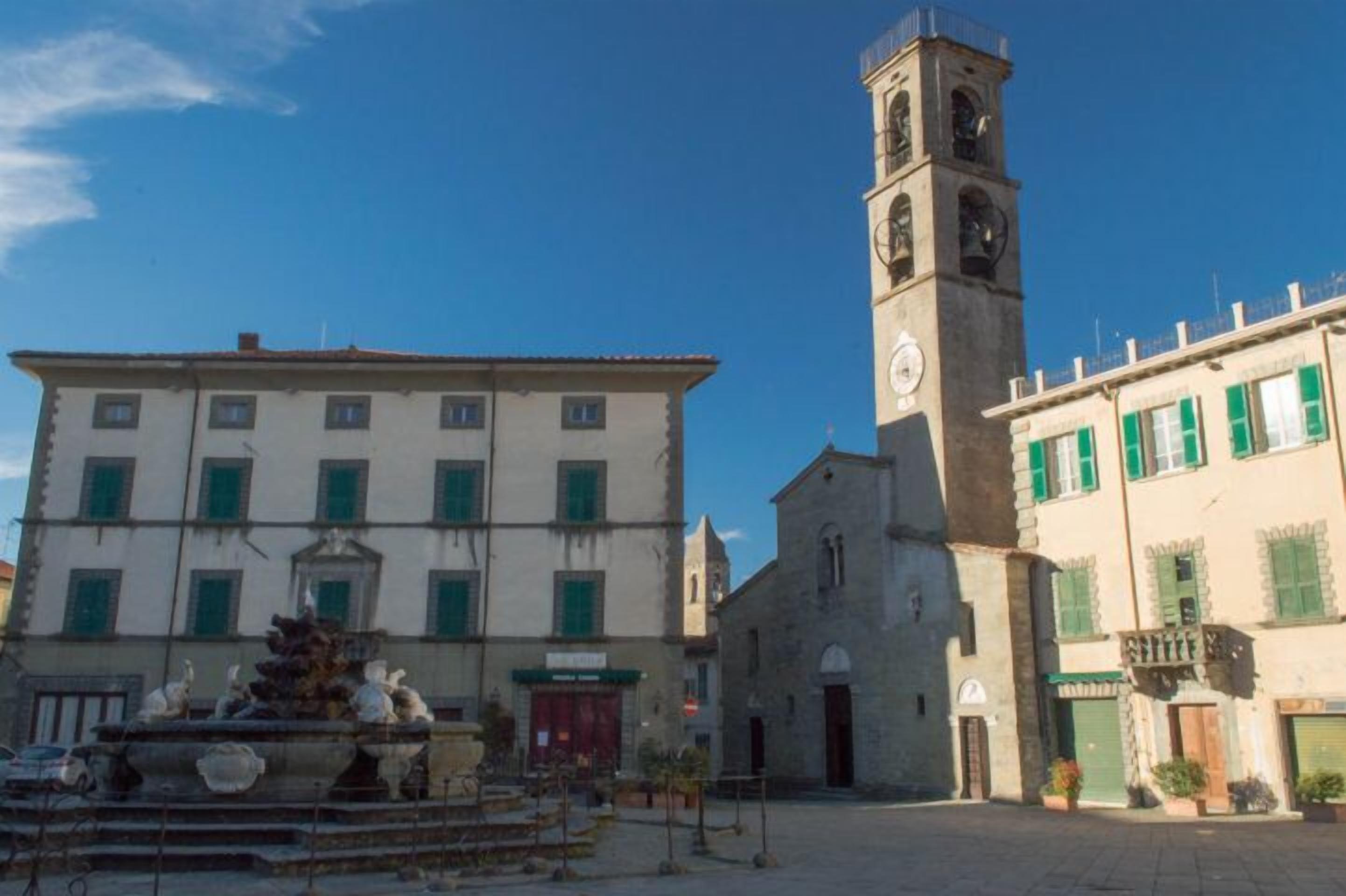A square with a fountain, a church with a bell tower, and a building with green shutters.