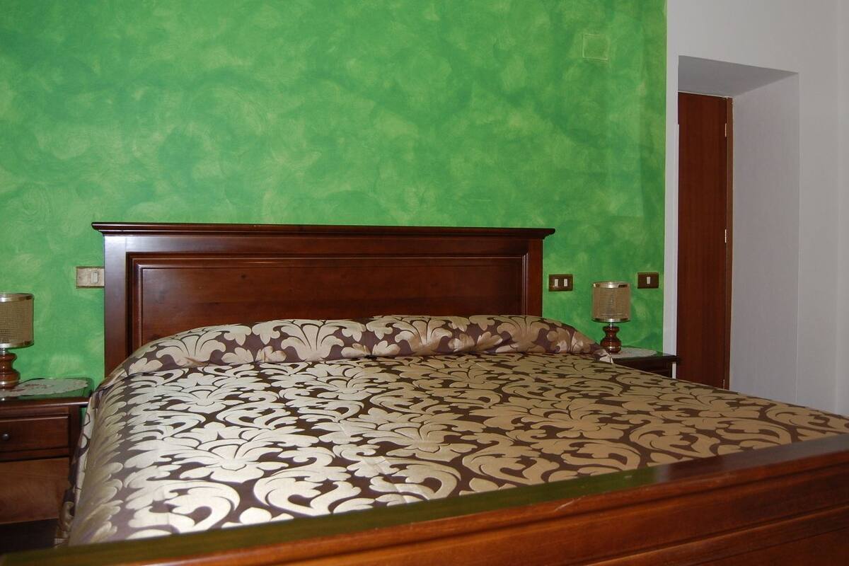 Ein Bett mit gemustertem Überwurf, ein Holz-Kopfende, ein Nachttisch mit Lampe und ein Stuhl in einem Zimmer mit grünen Wänden und einer Holztür.