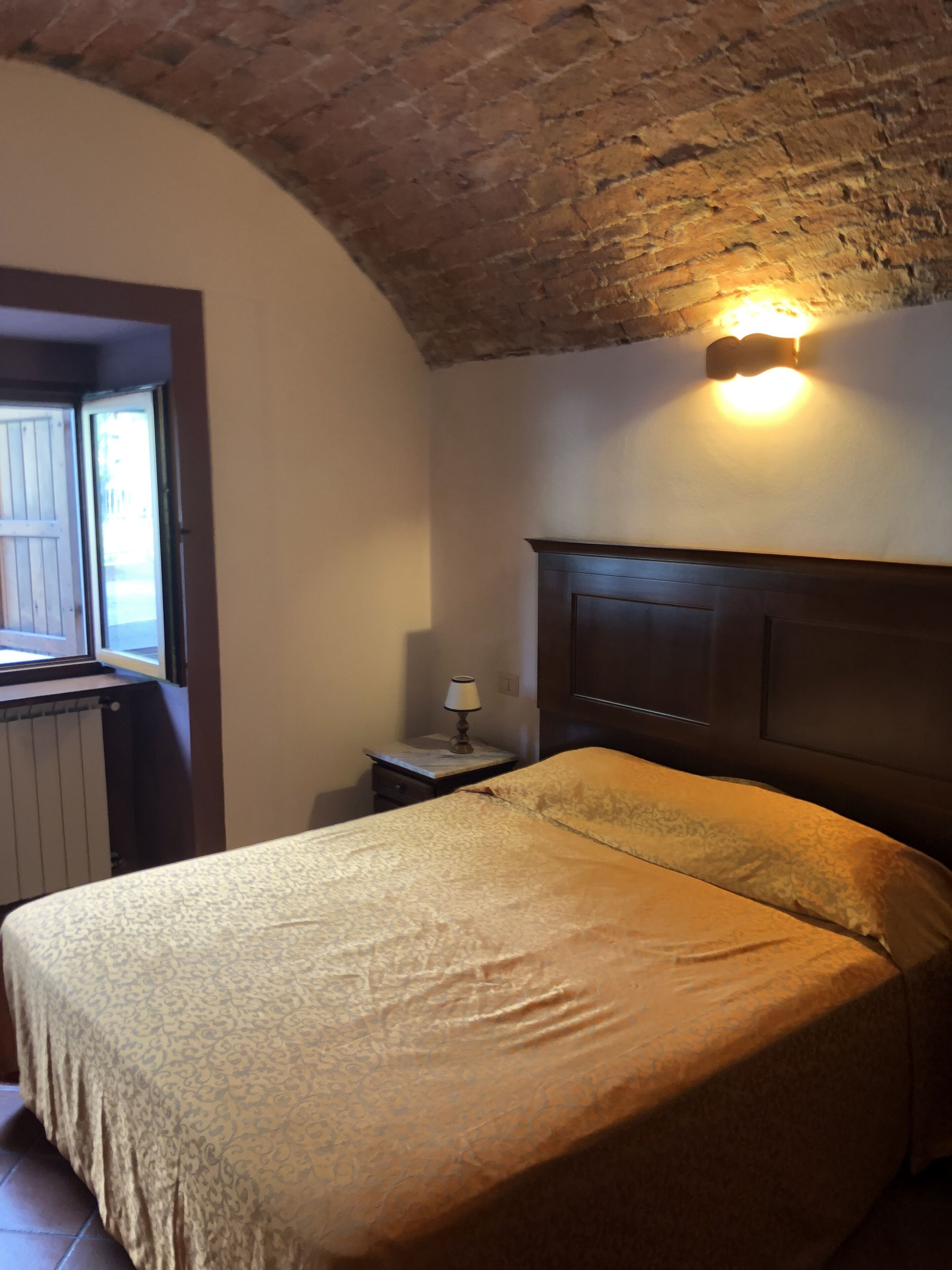 A bedroom with a bed, a nightstand, a lamp, a window, and a brick ceiling.