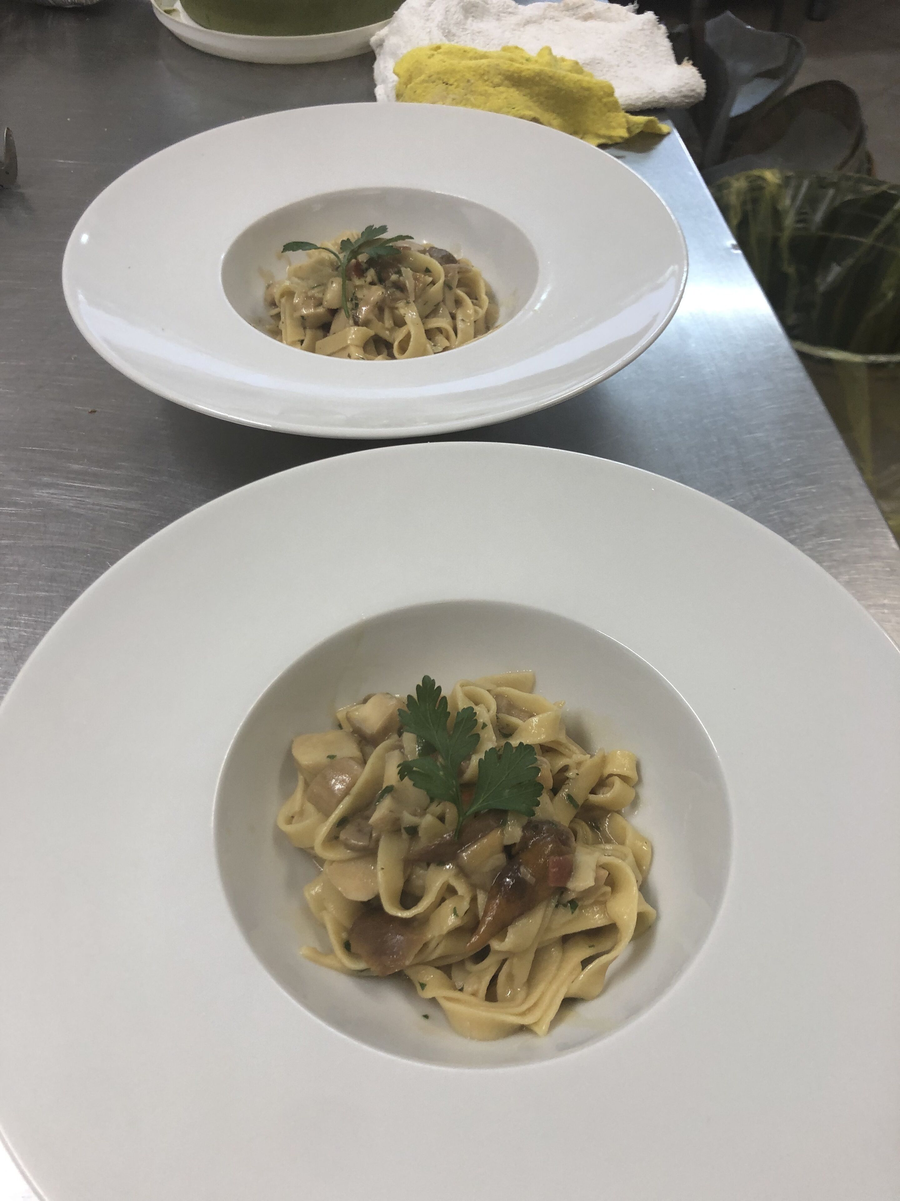 Two white plates of pasta with mushrooms and parsley on a stainless steel table.
