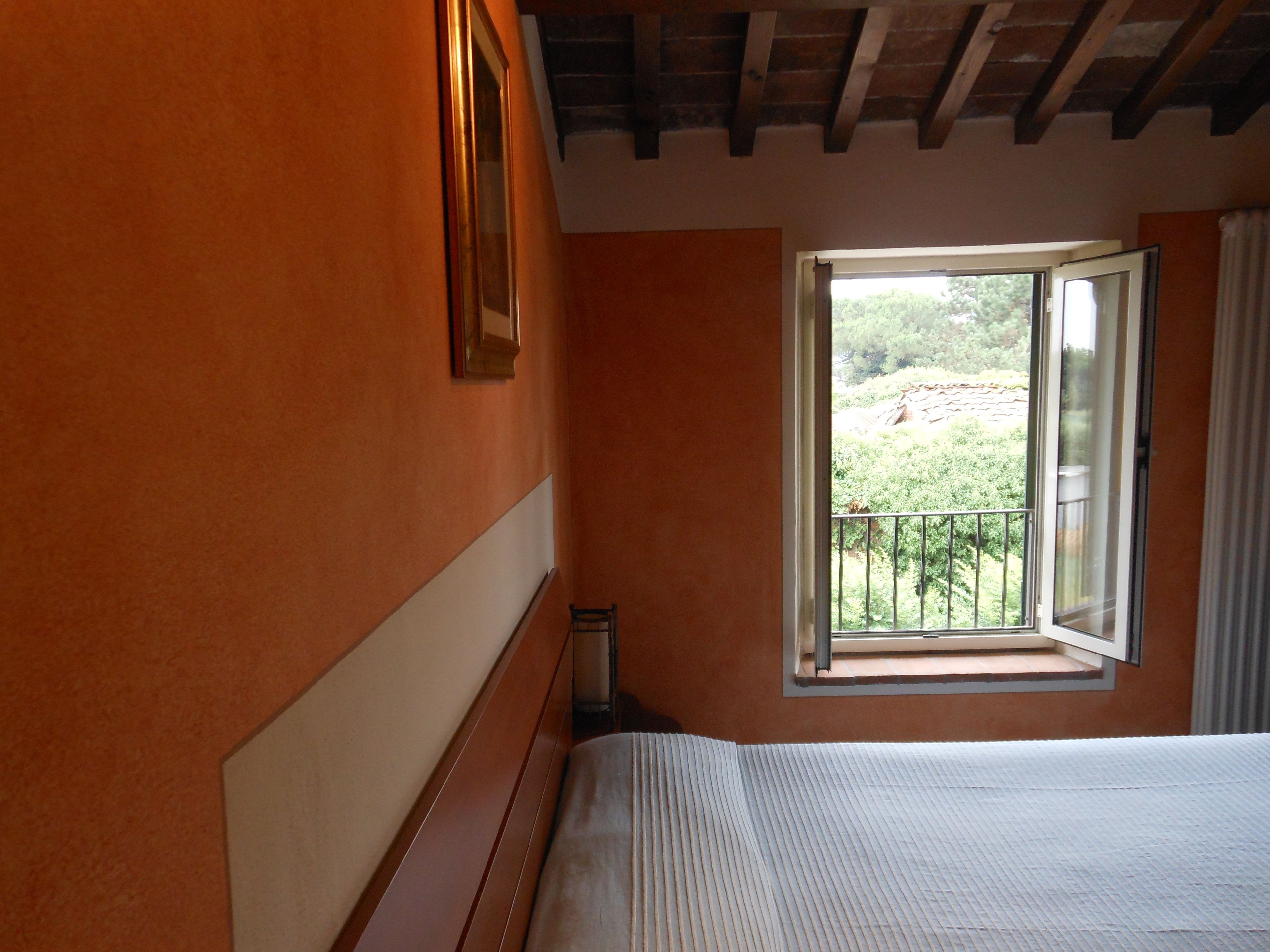 Ein Schlafzimmer mit großem Fenster, Holzbalken an der Decke und einem Bett mit weißem Kopfteil.