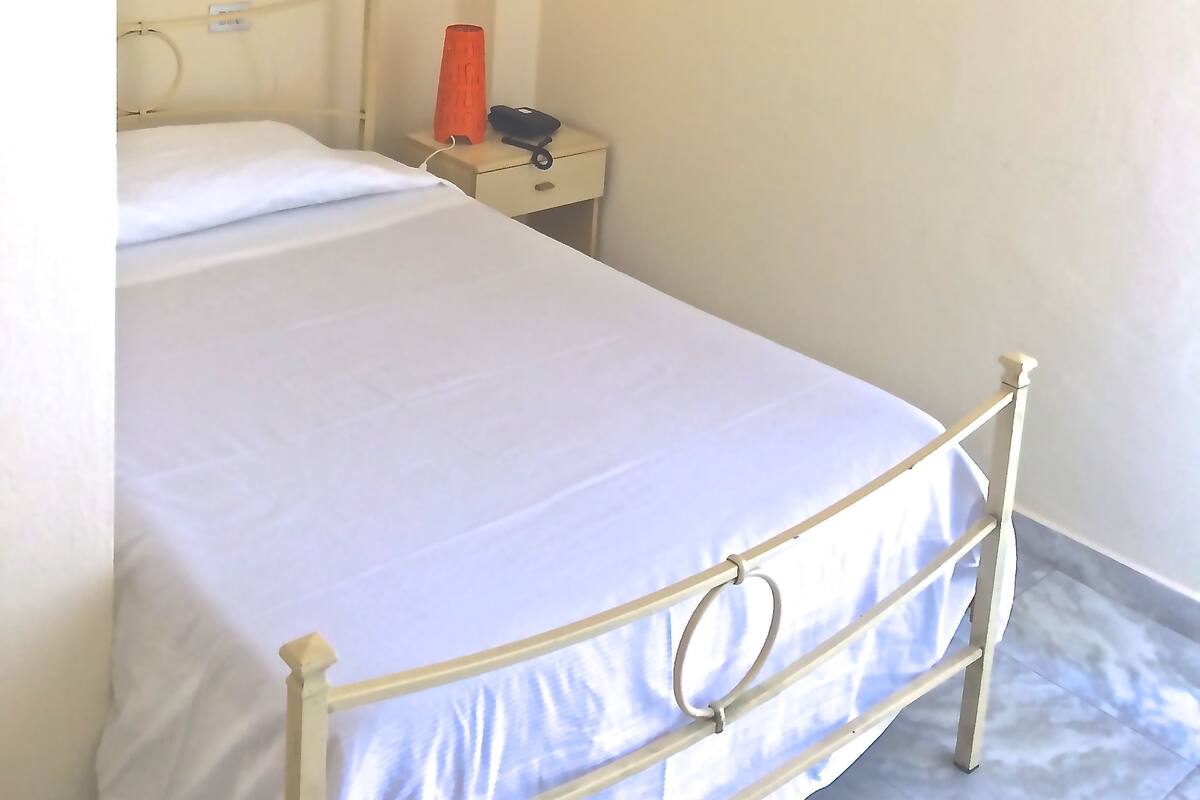 Een enkel bed met metalen frame, een nachtkastje met lamp en een airconditioning aan de muur.
