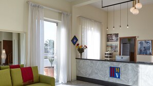 A hotel lobby with a green sofa, a reception desk, and a large window with curtains. - Hotel Mirage Versilia (Pietrasanta)