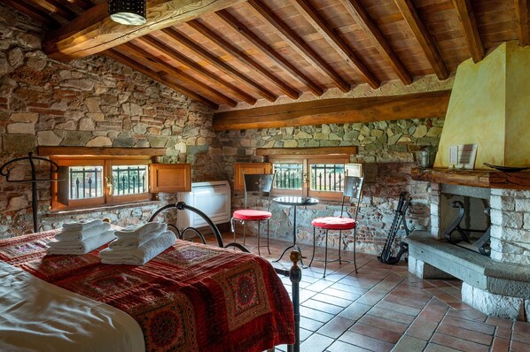 A rustic room with stone walls, wooden beams, a bed with a red blanket, two red stools, a stone bench, and a fireplace. - Villa Corte Degli Dei (Lucca)