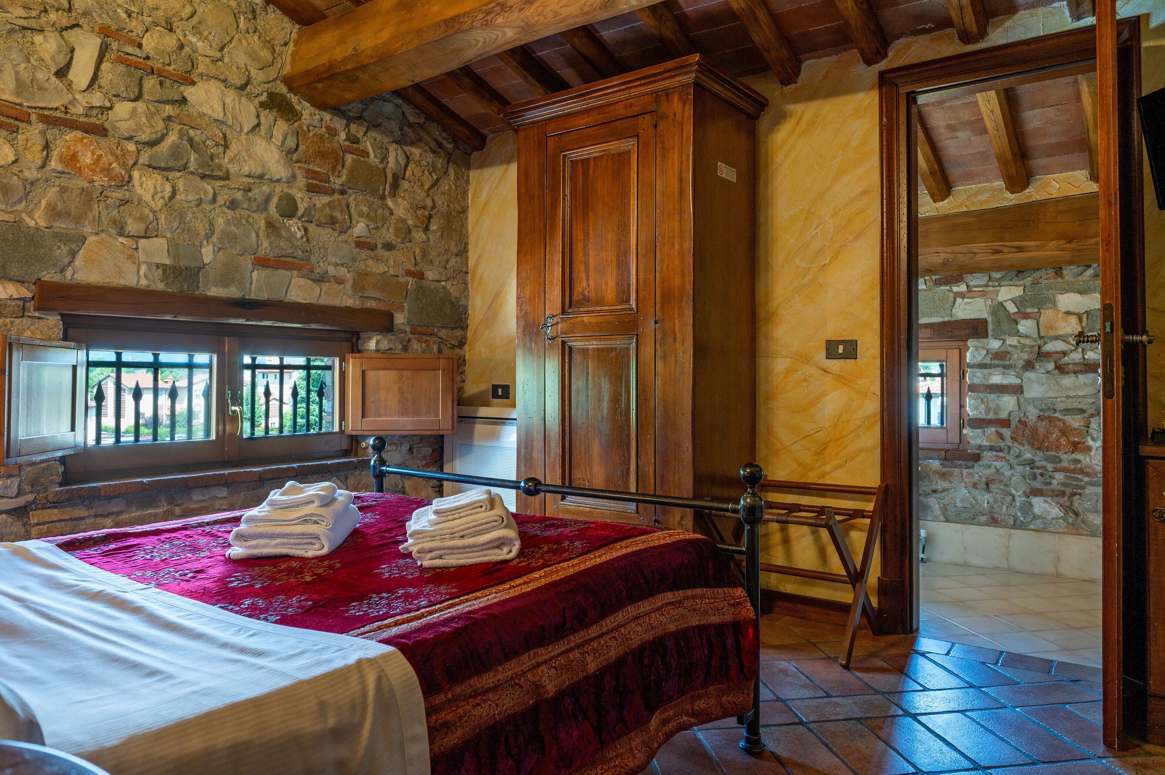 A room with a stone wall, wooden ceiling beams, a bed with a red bedspread, and a wooden wardrobe.