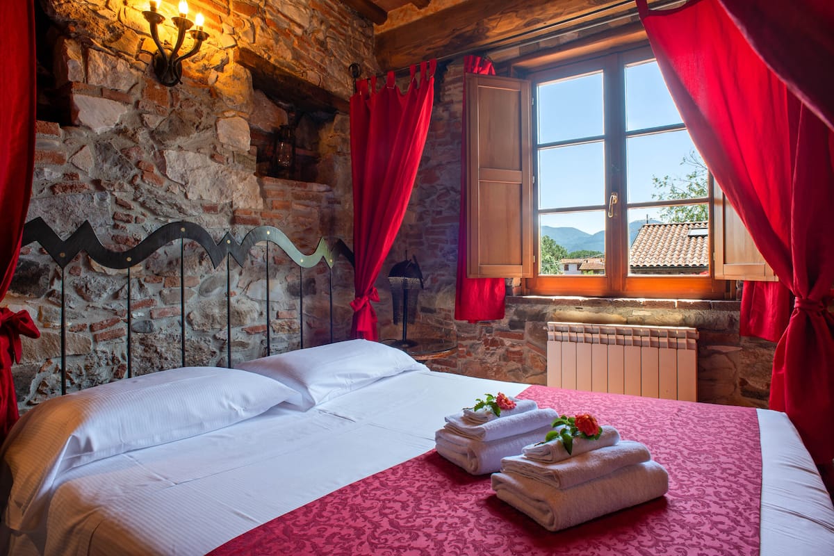 A room with a stone wall, a bed with white linens, red curtains, and a window with a view of mountains.