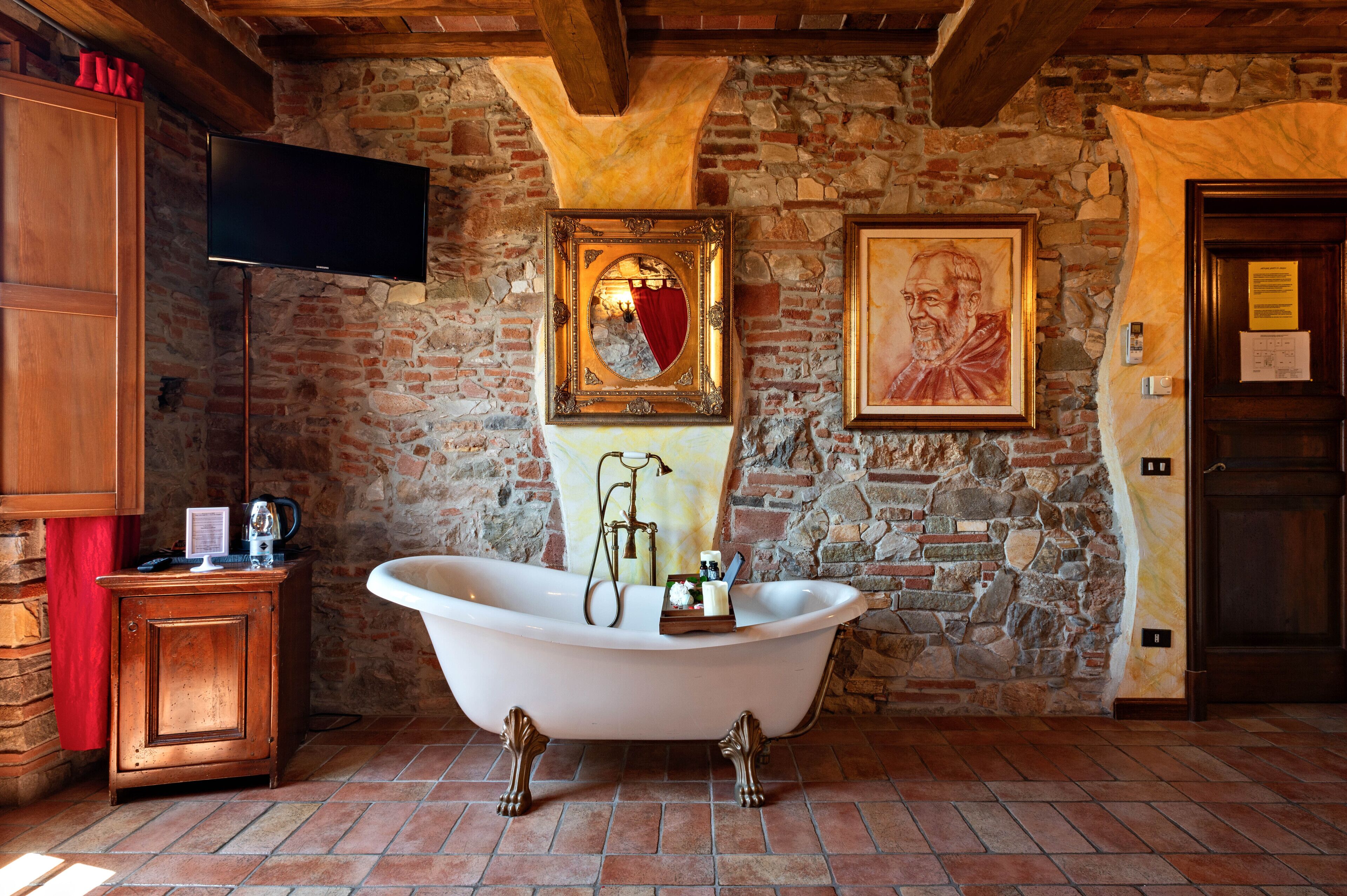 A rustic bathroom with a clawfoot bathtub, a wooden cabinet, a stone wall, and a mounted television.