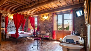 A rustic room with a stone wall, a wooden ceiling, a bed with red curtains, a table with chairs, and a bathtub. - Villa Corte Degli Dei (Lucca)