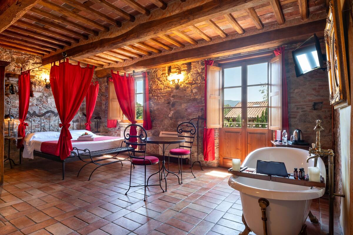 A rustic room with a stone wall, a wooden ceiling, a bed with red curtains, a table with chairs, and a bathtub.