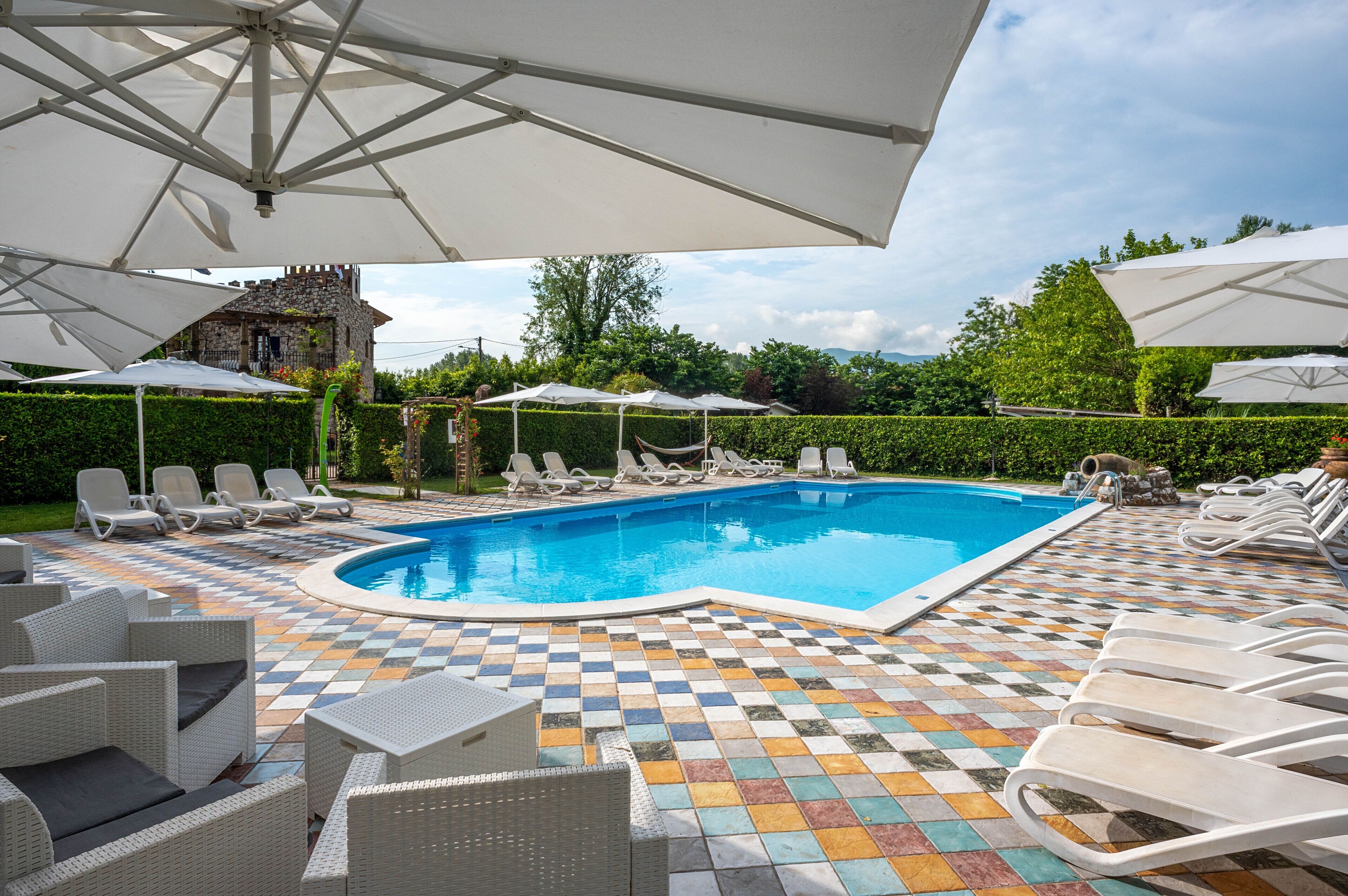 A pool area with lounge chairs and umbrellas, surrounded by a checkered tile floor and greenery.