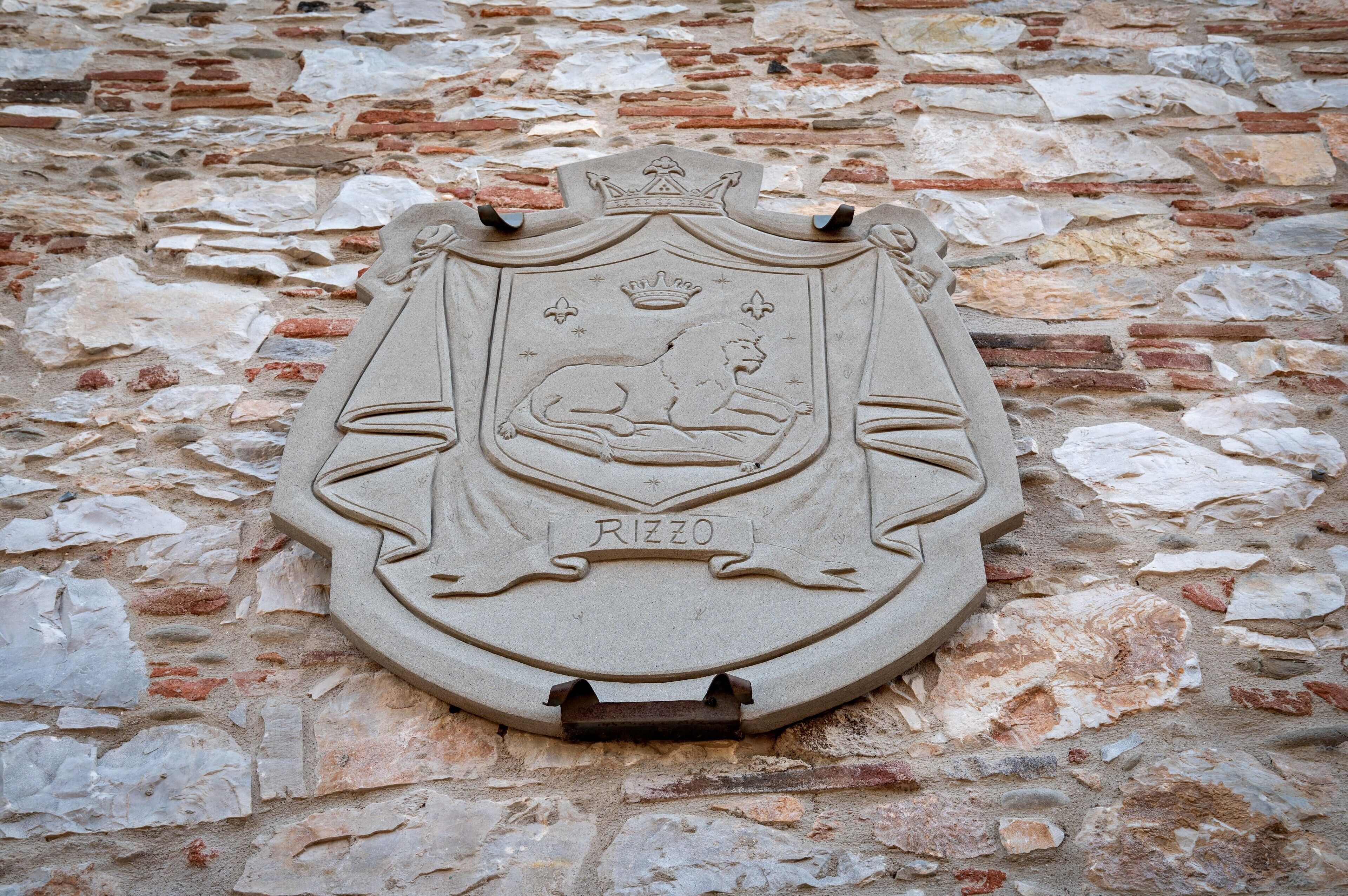 A stone coat of arms with a lion and the word "RIZZO" on a brick wall.