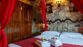 Habitación de hotel con pared de piedra, armario de madera, cortinas rojas, cama con ropa blanca y colcha estampada de color rojo.