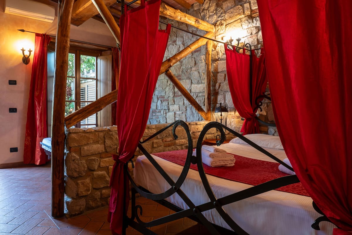A stone-walled bedroom with a wooden bed frame, red curtains, and a stone fireplace.